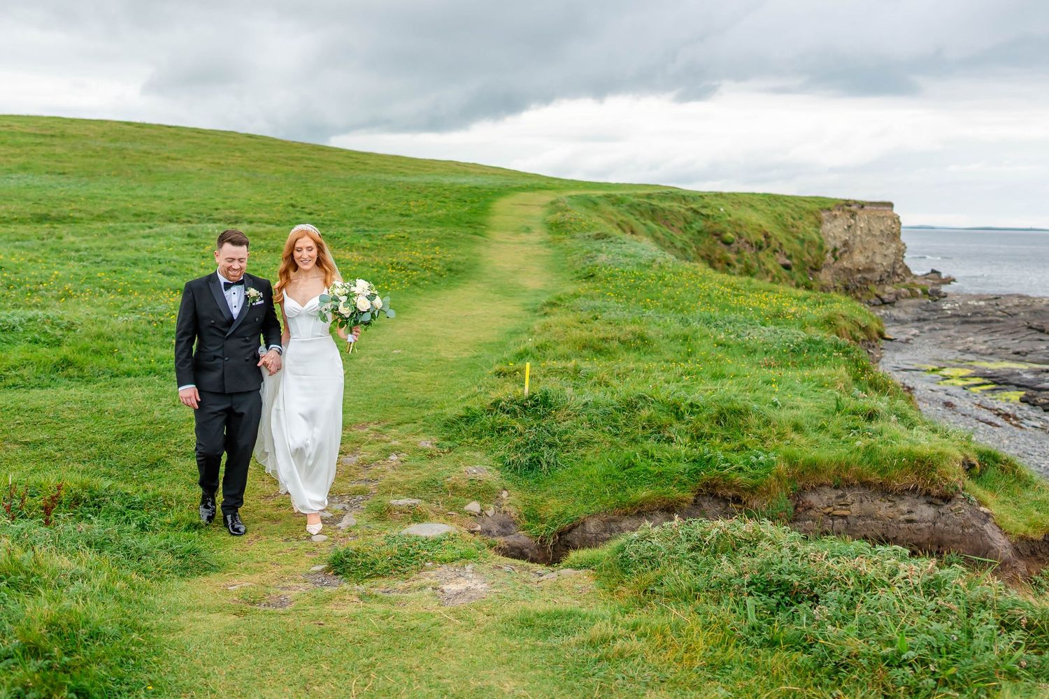 Wedding couple explores the rugged coastline with dramatic cliffs and green fields in the background.