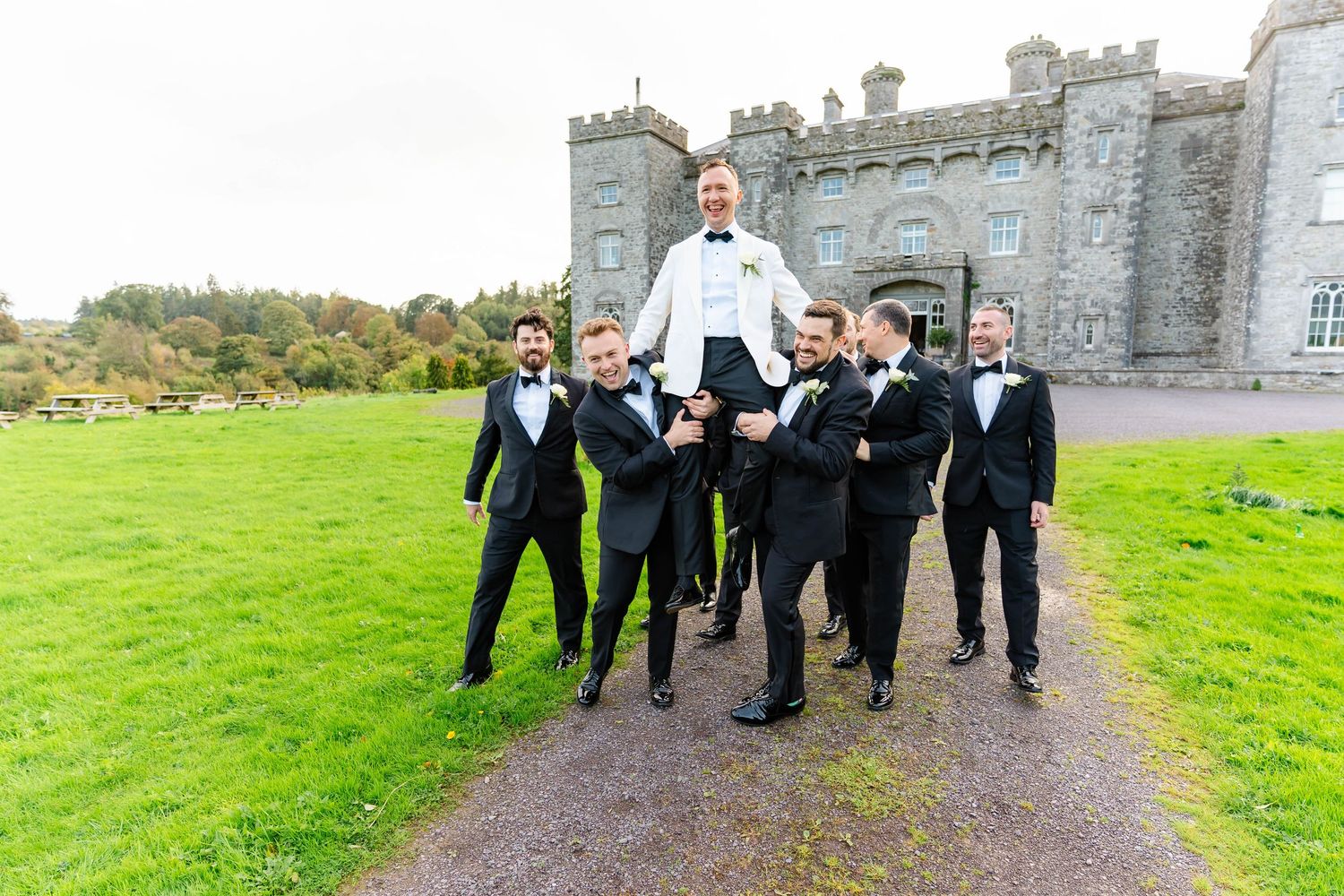 A group in formal wedding attire joyfully runs through the green grounds of a castle estate.