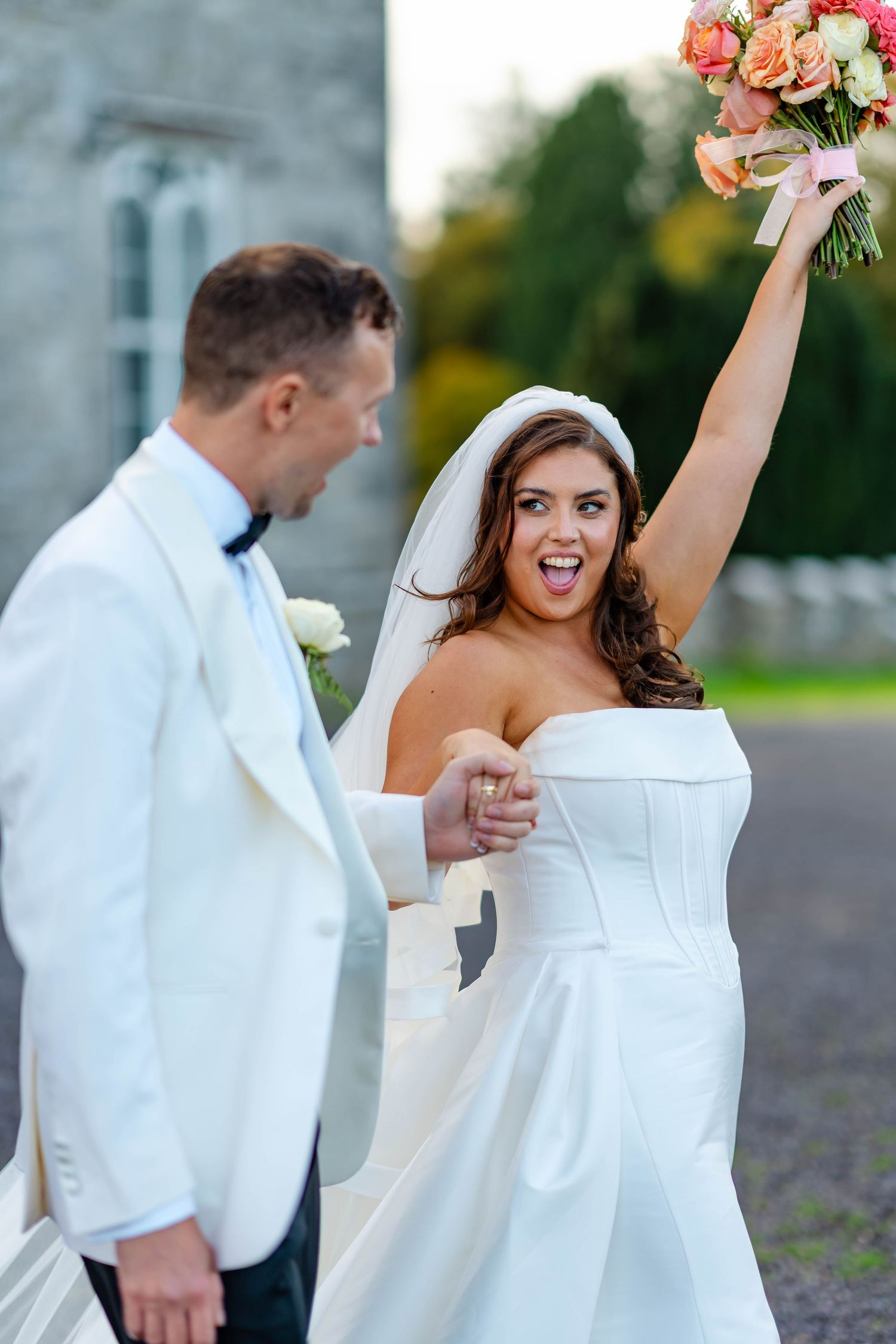 A joyful bride in a white strapless gown celebrates with raised arms while holding a coral and white bouquet.