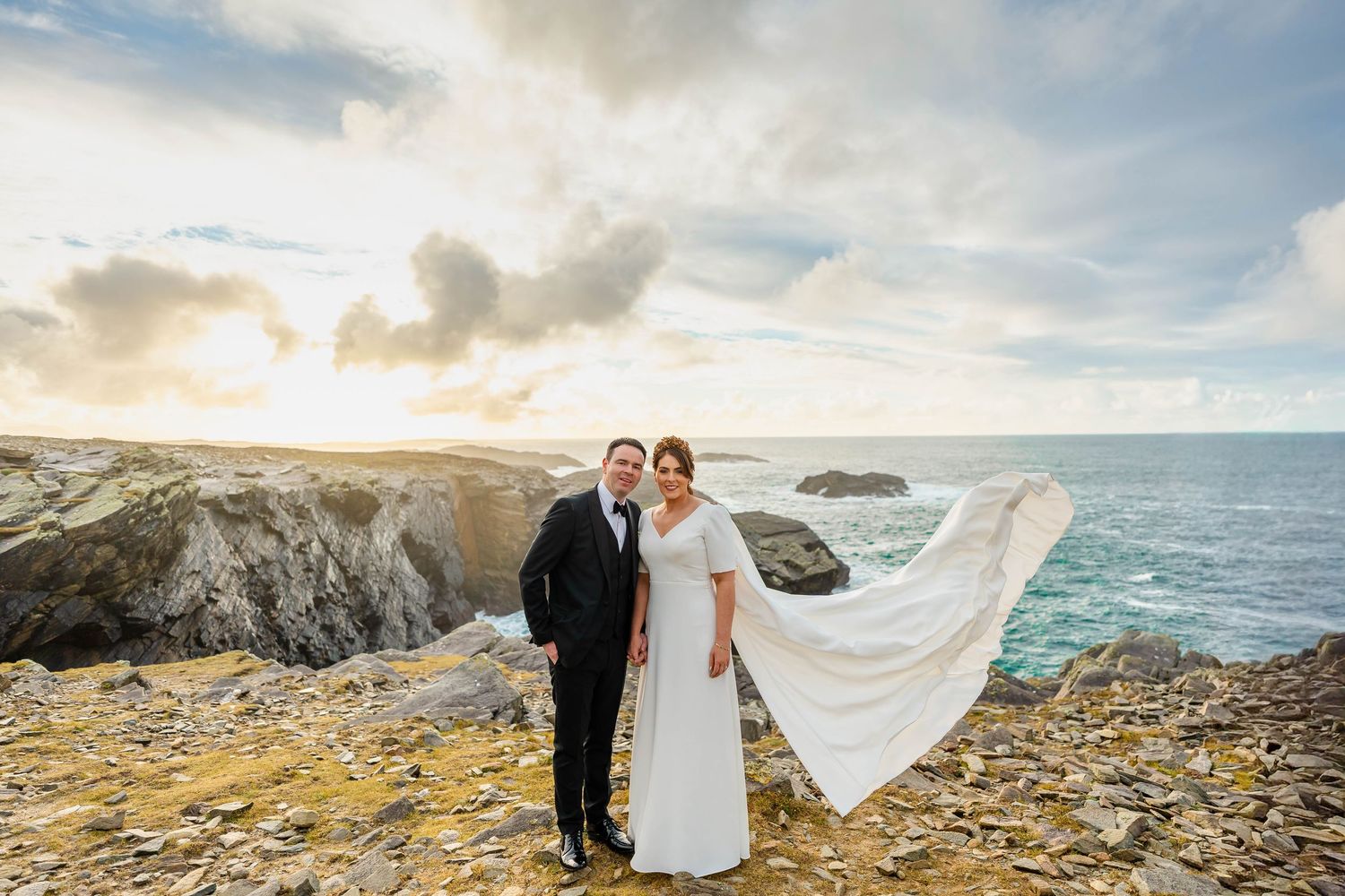 Wedding couple on cliff edge with flowing veil catching wind against dramatic coastal sunset.