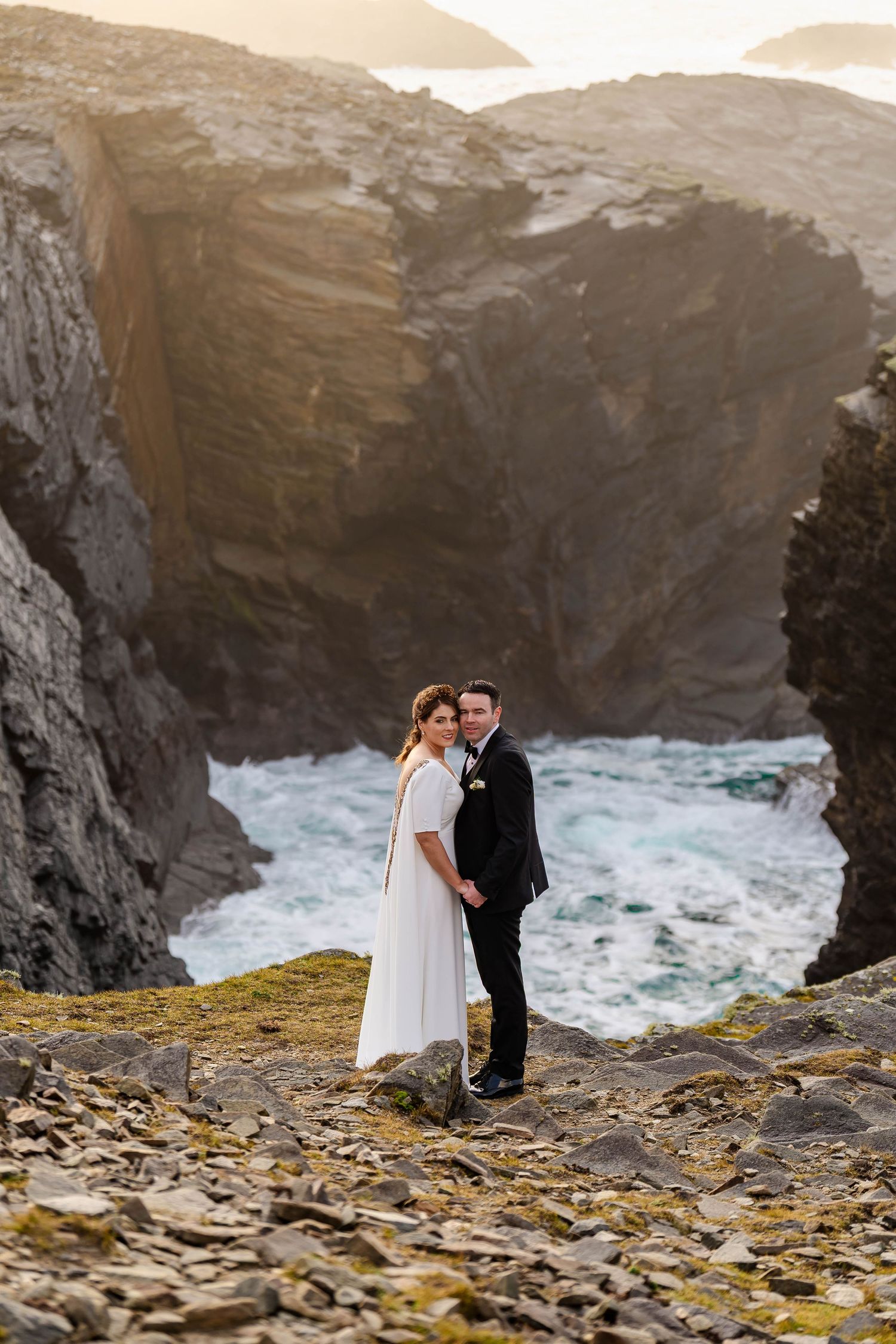 Wedding couple poses against dramatic coastal landscape with rocky cliffs and churning ocean.