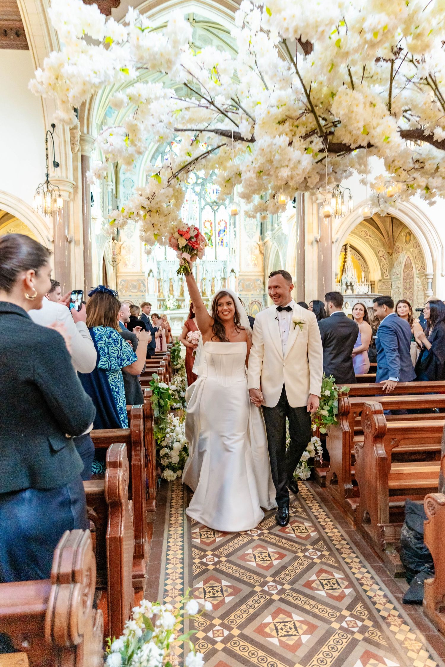 Wedding guests gather in an ornate church with decorative floor tiles and floral arrangements during a ceremony.