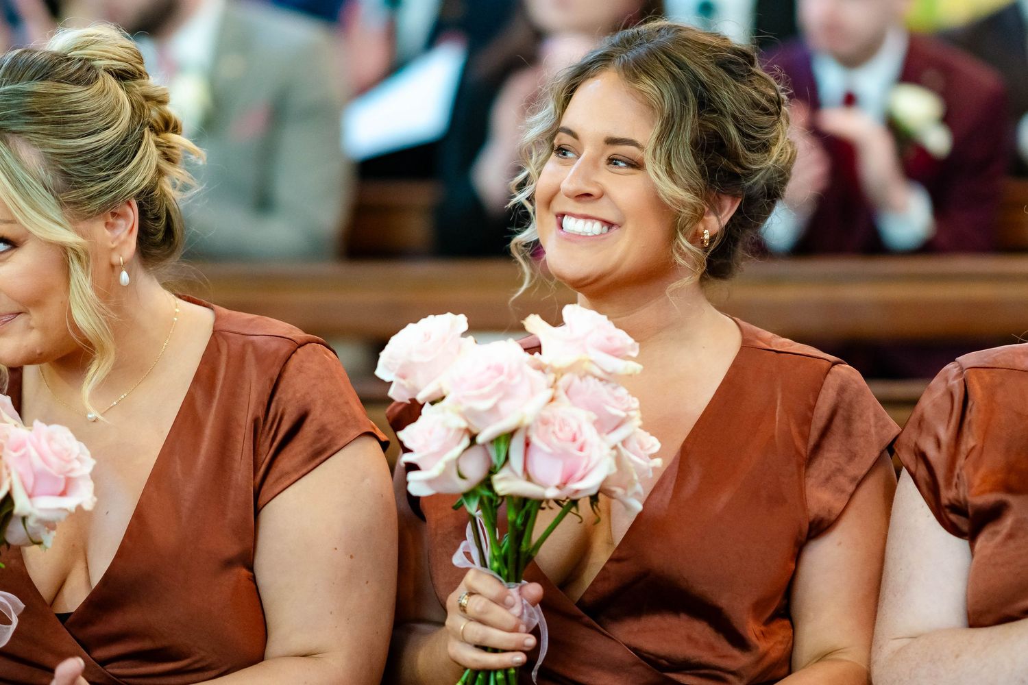 Bridesmaids in rust-colored dresses smile while holding pink peony bouquets during wedding ceremony.