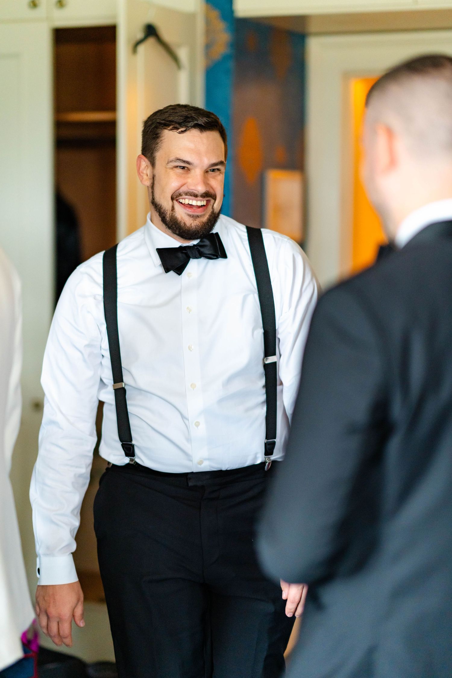 Groomsman in white dress shirt and black suspenders smiles during wedding preparations.