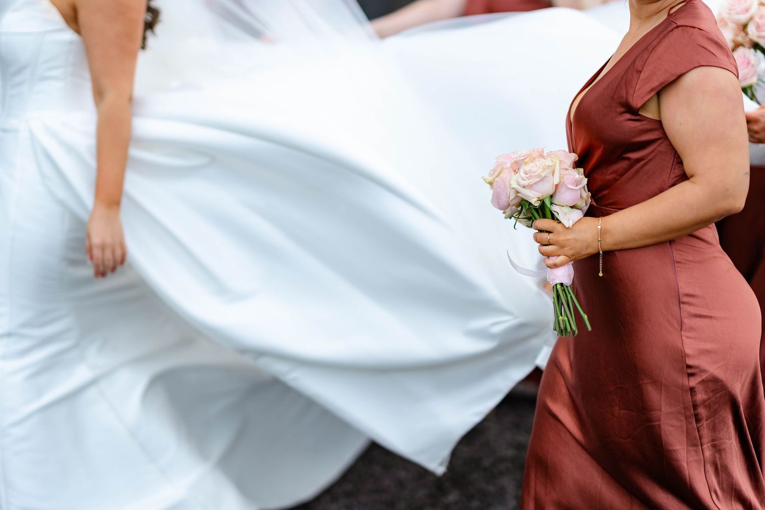 A person in a flowing white dress and another in a dusty rose gown hold a bouquet of pink flowers at a wedding.