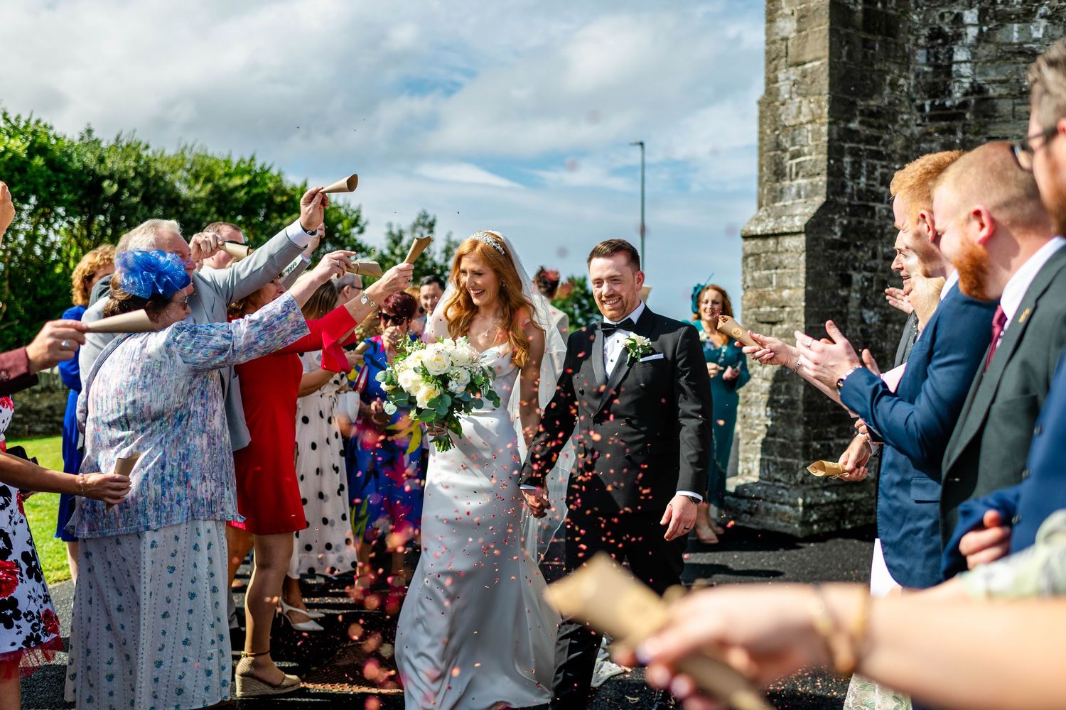 Wedding guests throwing confetti at newlyweds outside stone church on sunny day.
