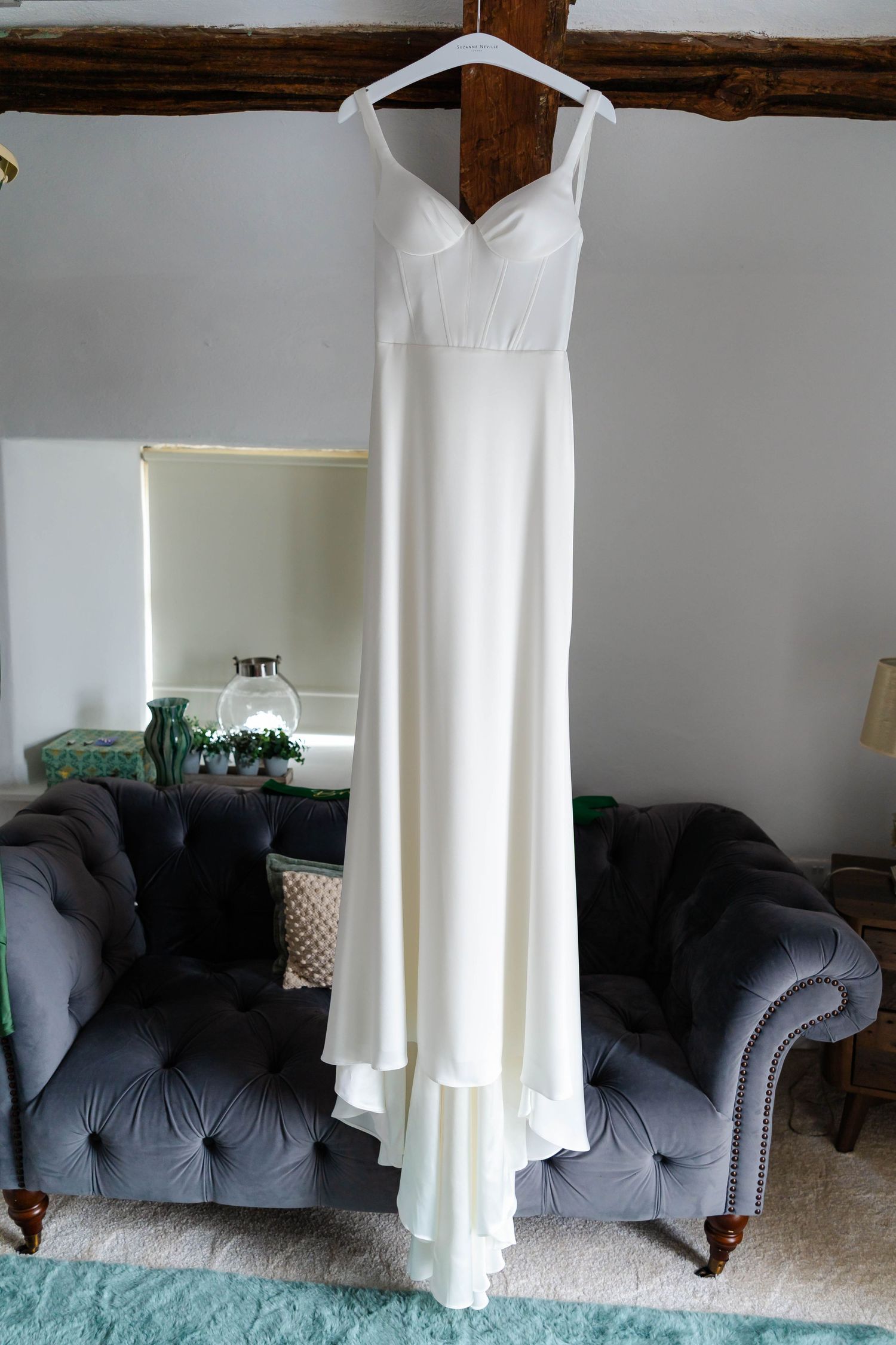 White wedding dress hanging from wooden beam against grey couch in room.