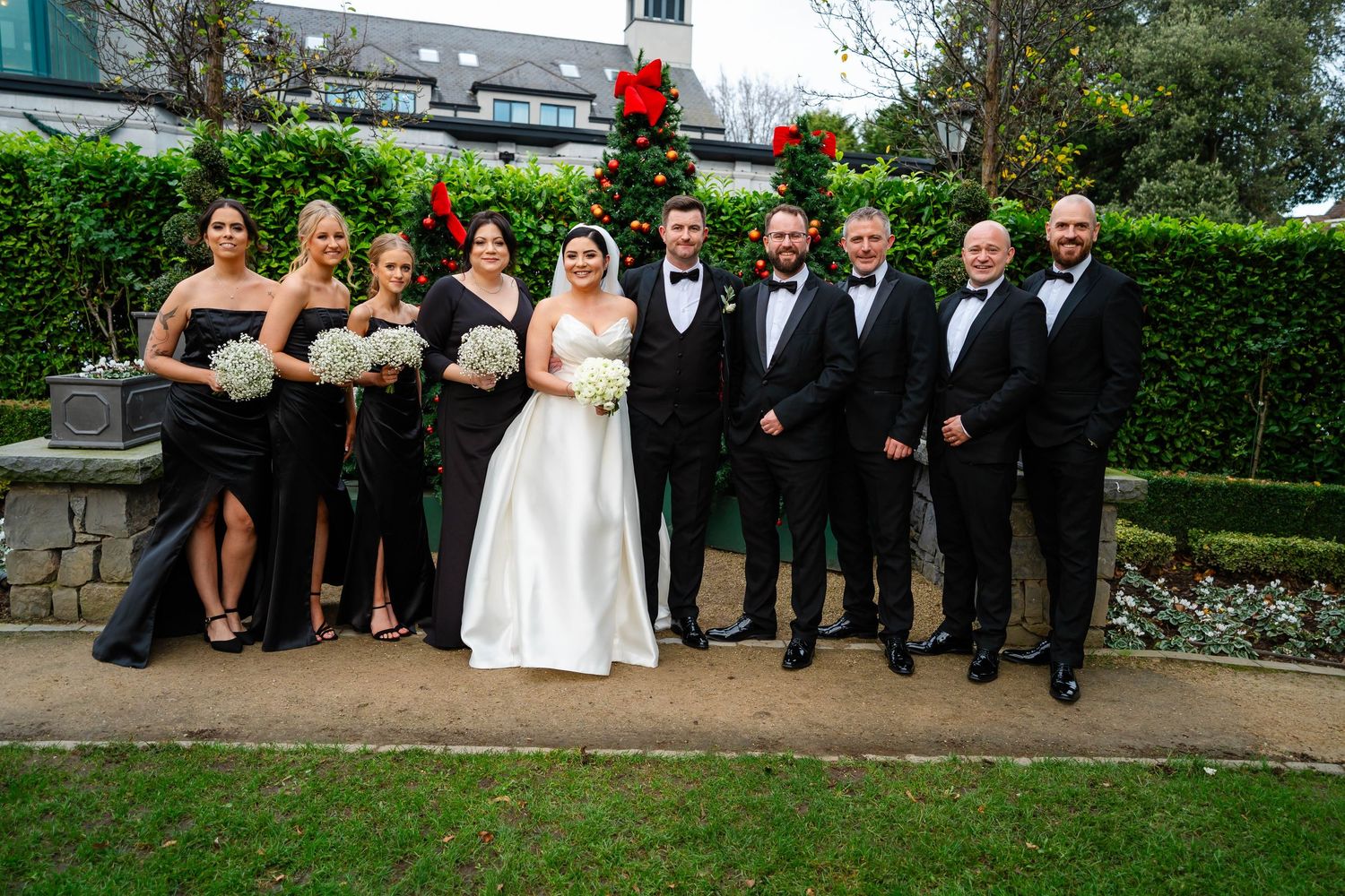 Wedding party in formal attire poses outdoors with bridesmaids in black and groomsmen in tuxedos.