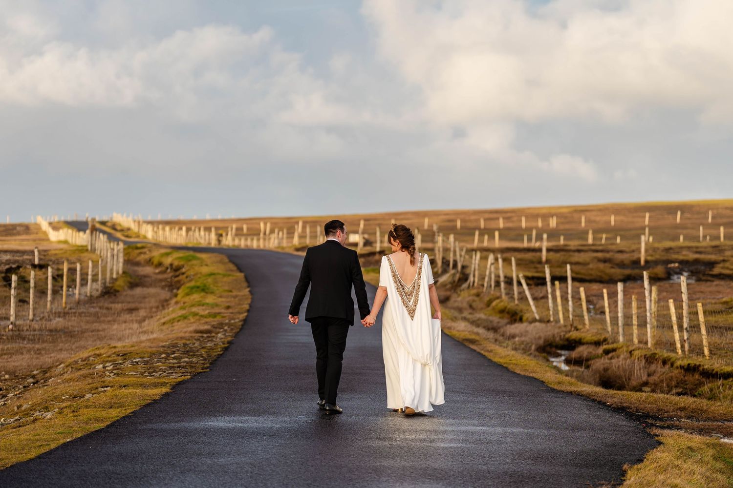 Newlyweds stroll down a winding country road at dusk with fence posts lining the path.