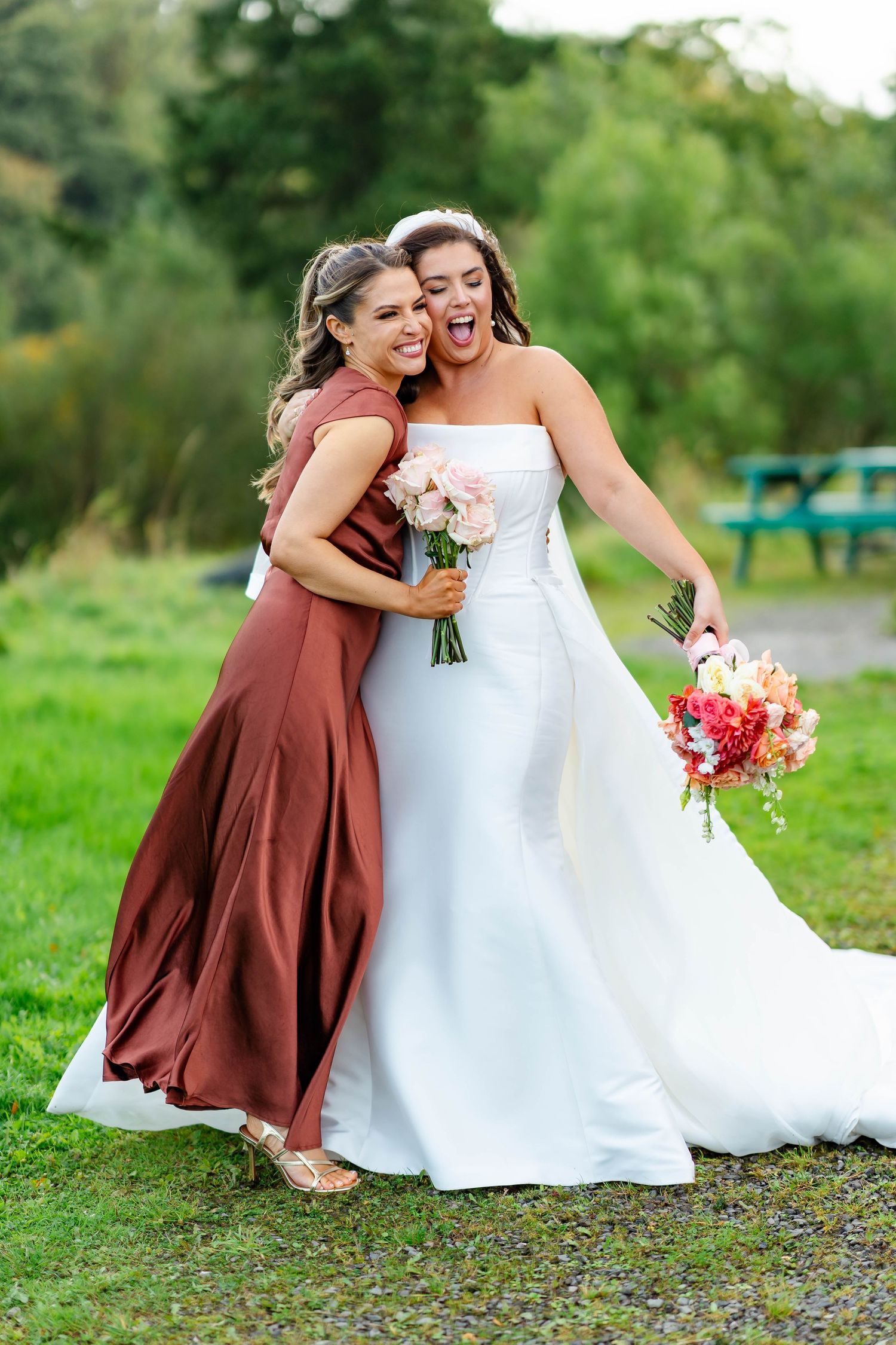 Two people in brown and white dresses share an intimate embrace outdoors in a lush green setting.