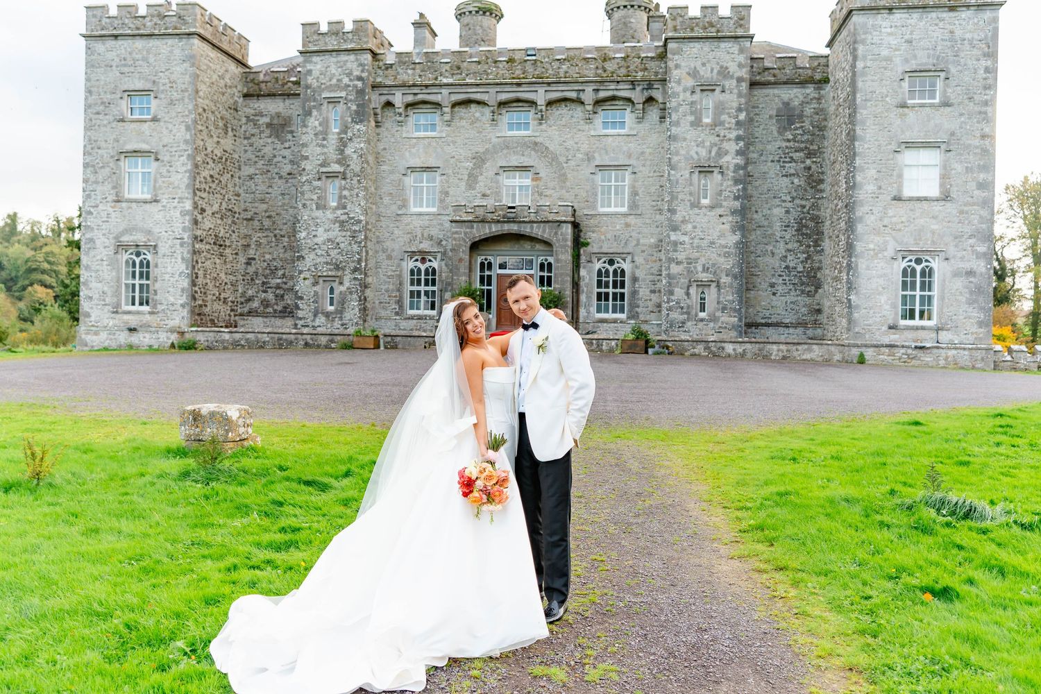 A couple in wedding attire poses in front of a historic stone castle with green grounds in Ireland.