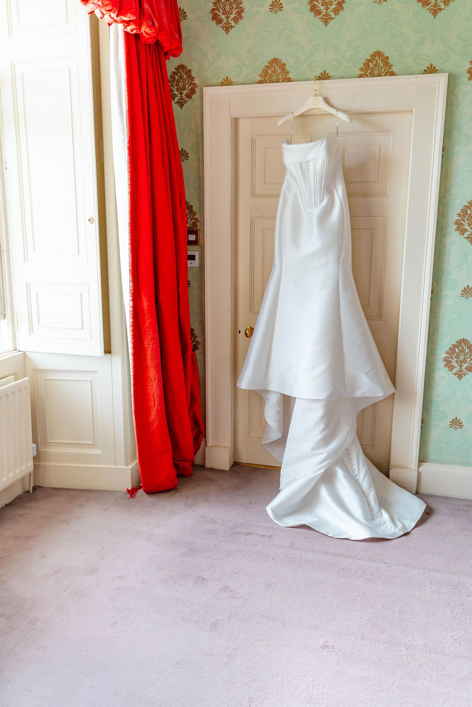 A white wedding dress displayed in an elegant room with mint green damask wallpaper and red curtains.