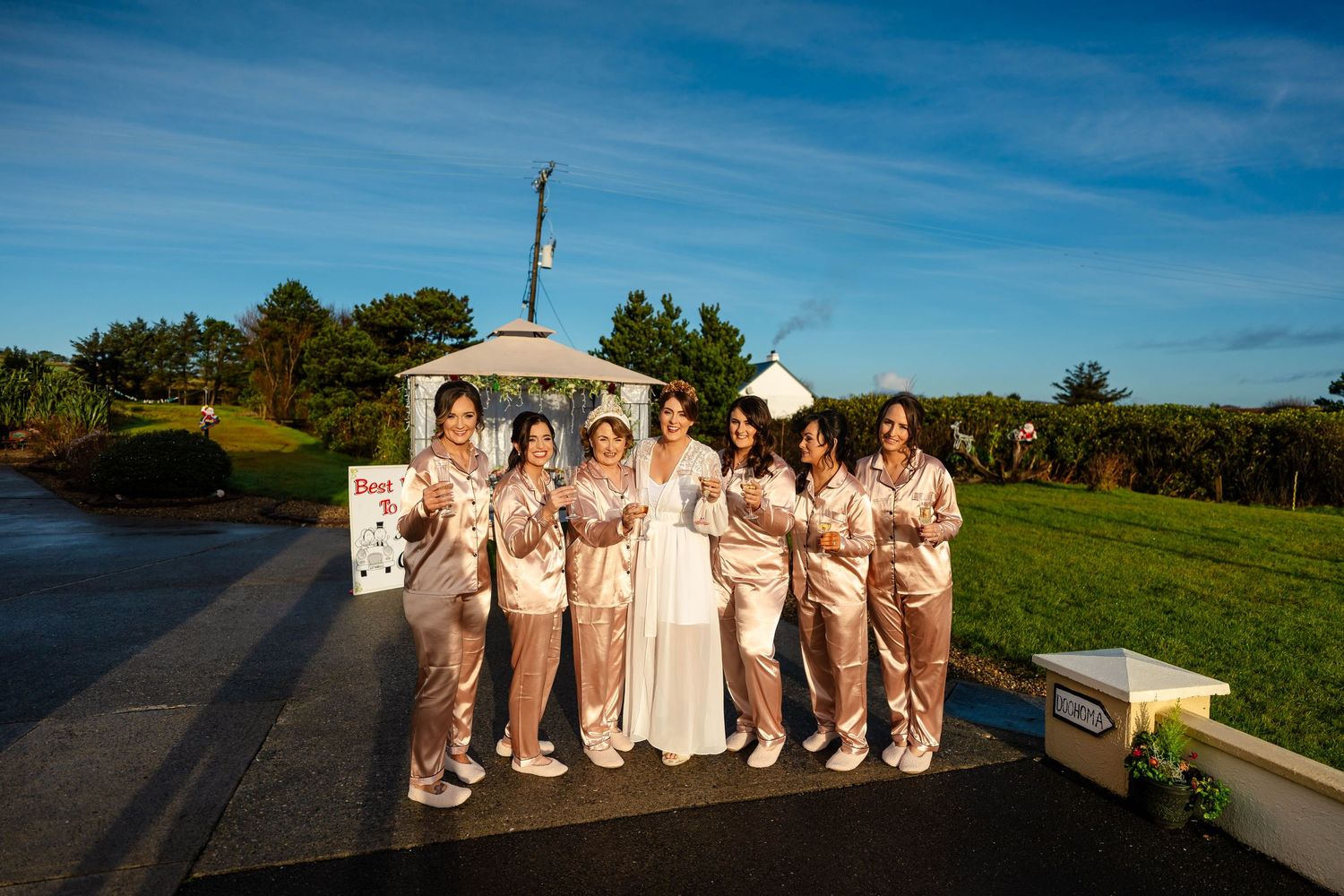 Wedding party members gathered outdoors under bright blue skies near a white building.