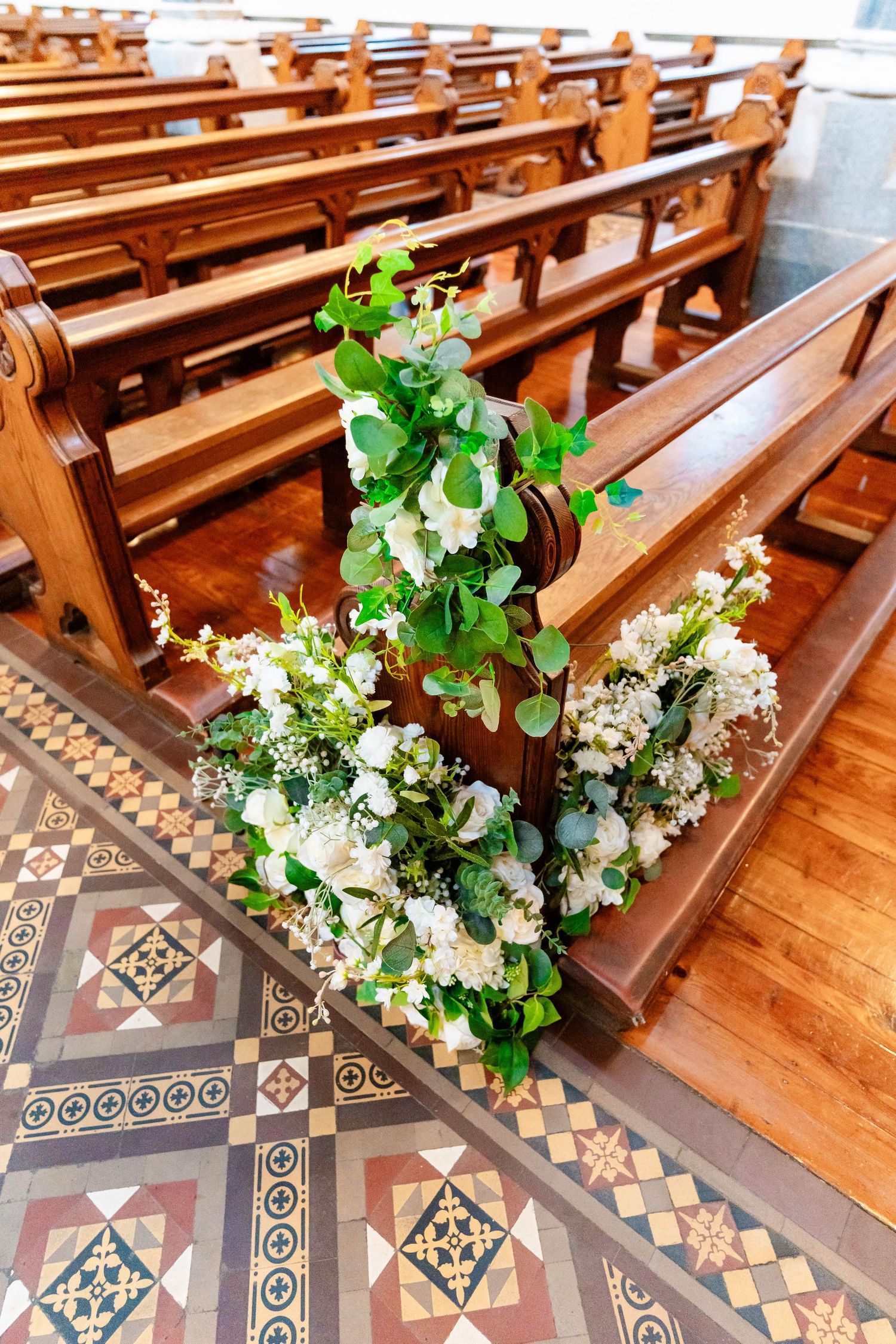 Church pews decorated with elegant white floral arrangements and greenery along the aisle in a historic chapel with ornate floors.