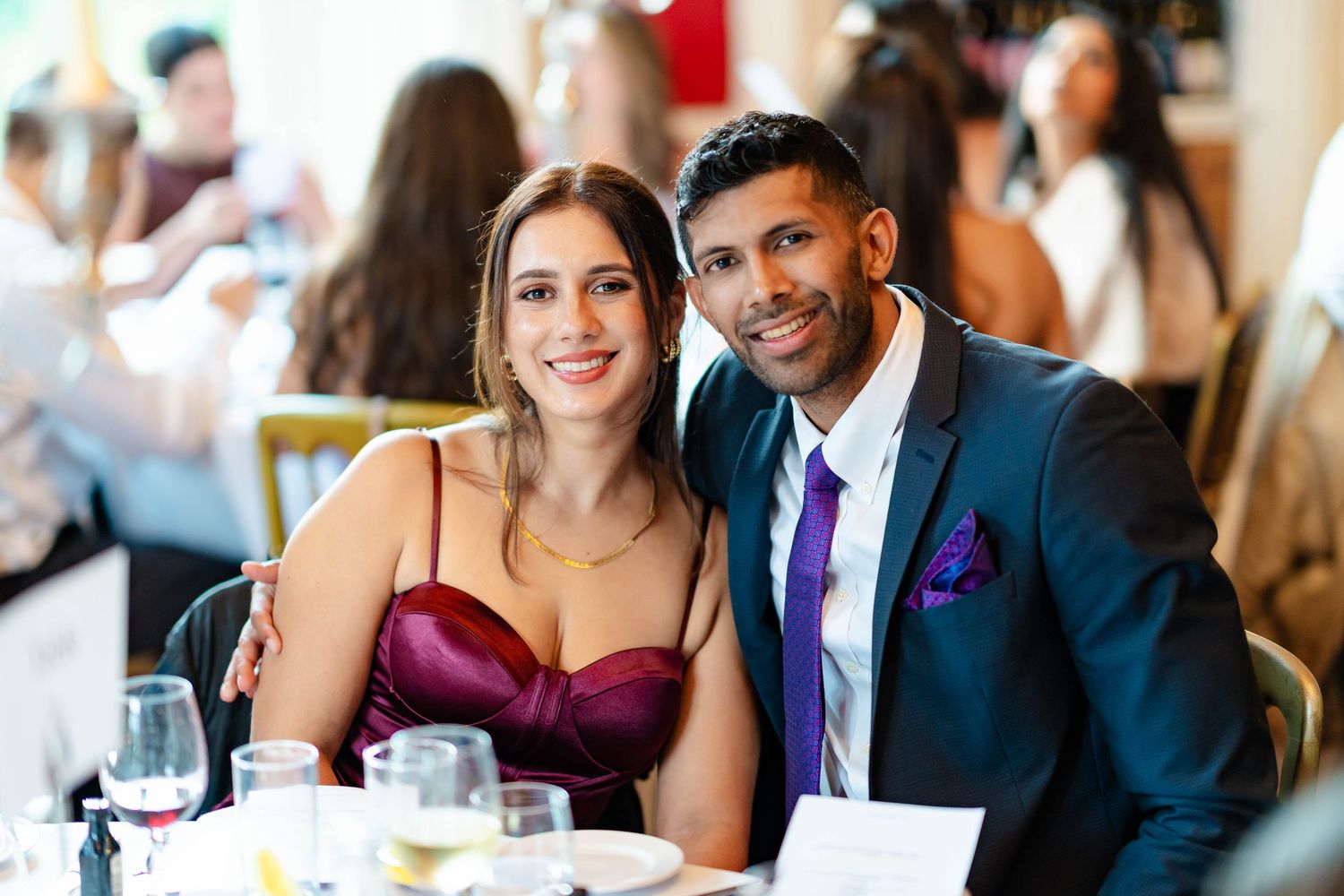 Wedding guests in formal attire smile and chat while seated at reception tables decorated with elegant place settings.
