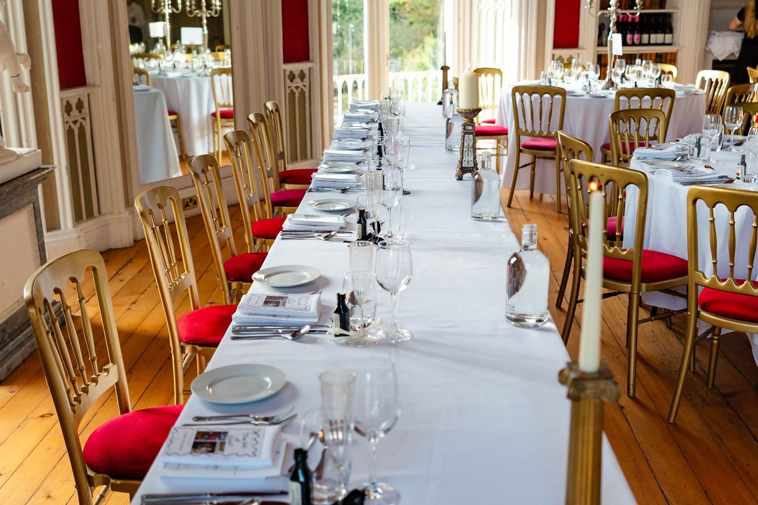 Elegant dining room interior with long white-clothed tables, red cushioned chairs, and natural light streaming through windows.