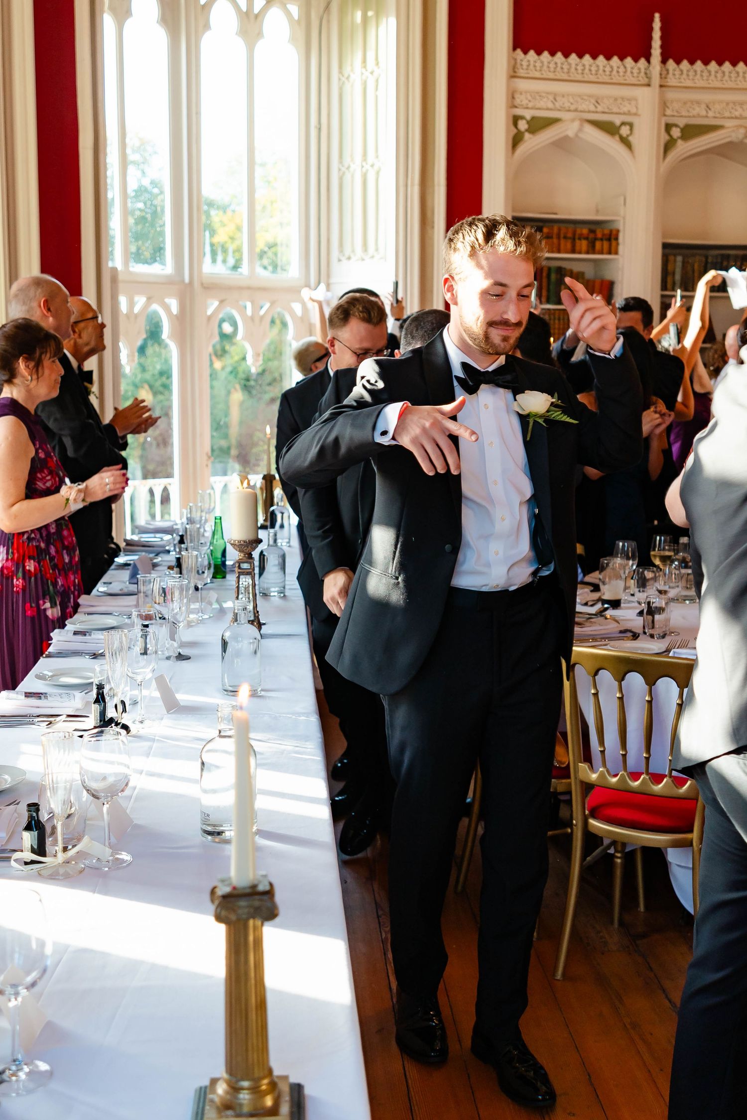 Wedding guests in formal attire dancing in an elegant reception hall with white-clothed tables and candle centerpieces.