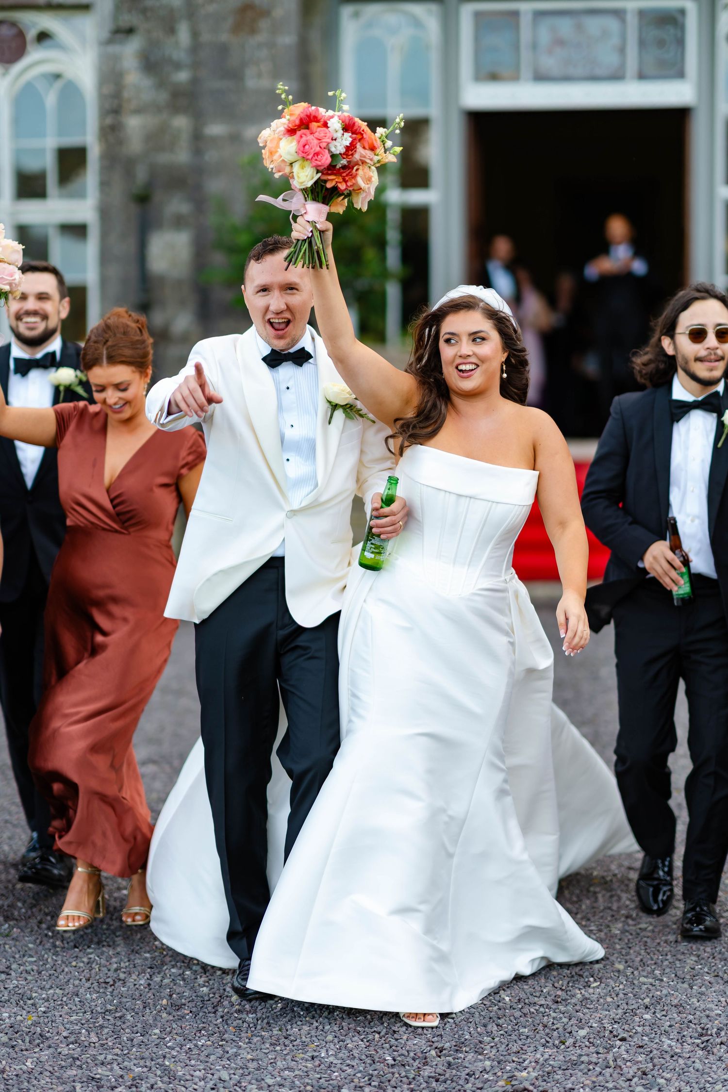 Wedding party in formal attire runs joyfully down a gravel path outside a stone building.