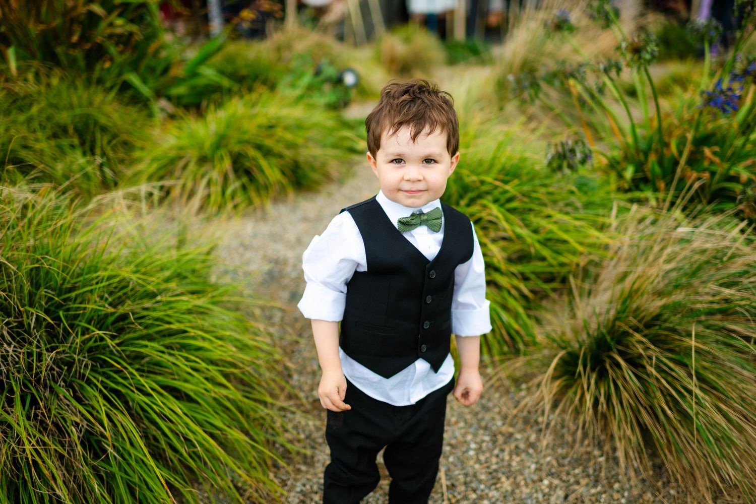 Young child in formal vest and bowtie stands among tall grass outdoors.
