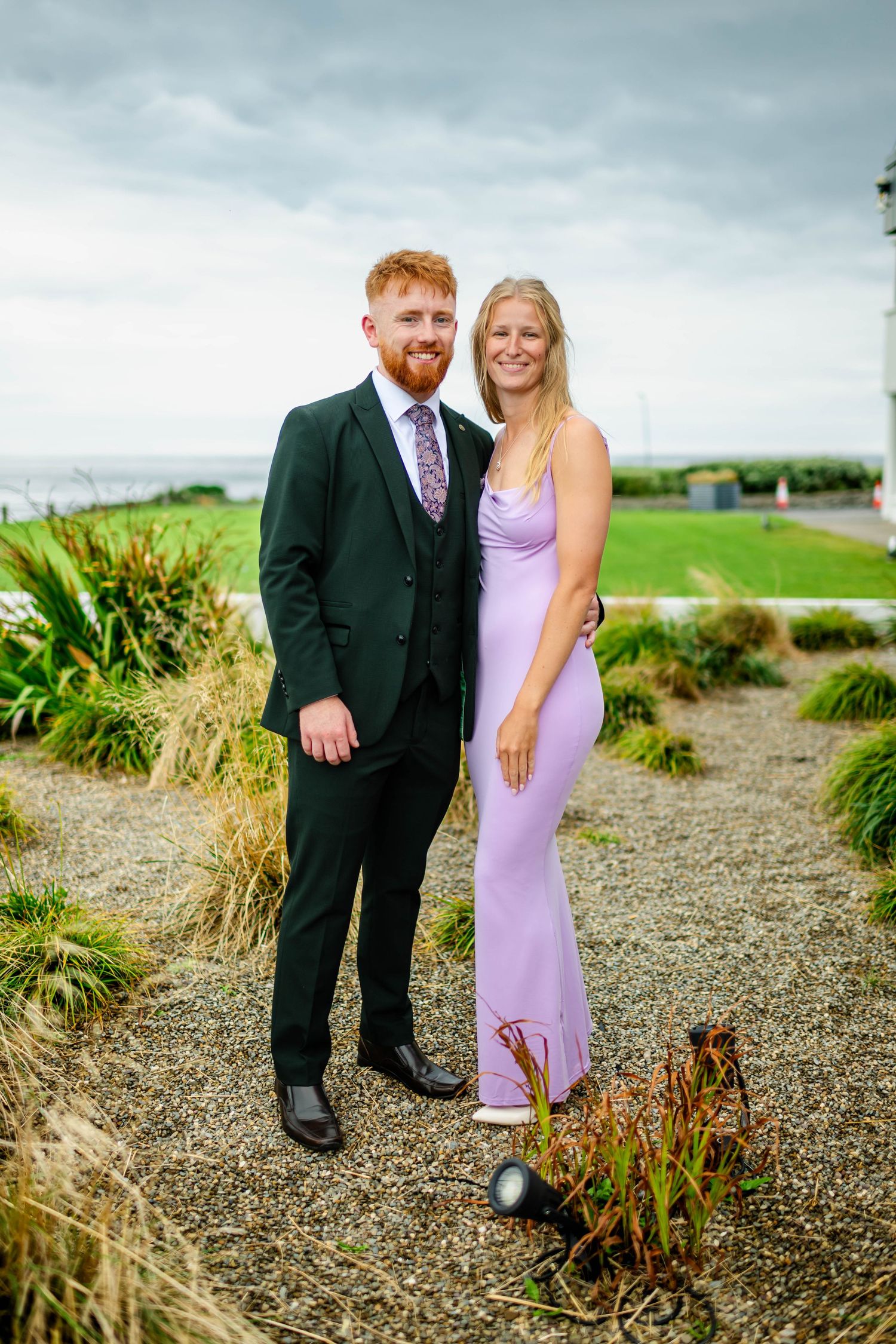 A couple poses outdoors in formal attire against a coastal landscape.