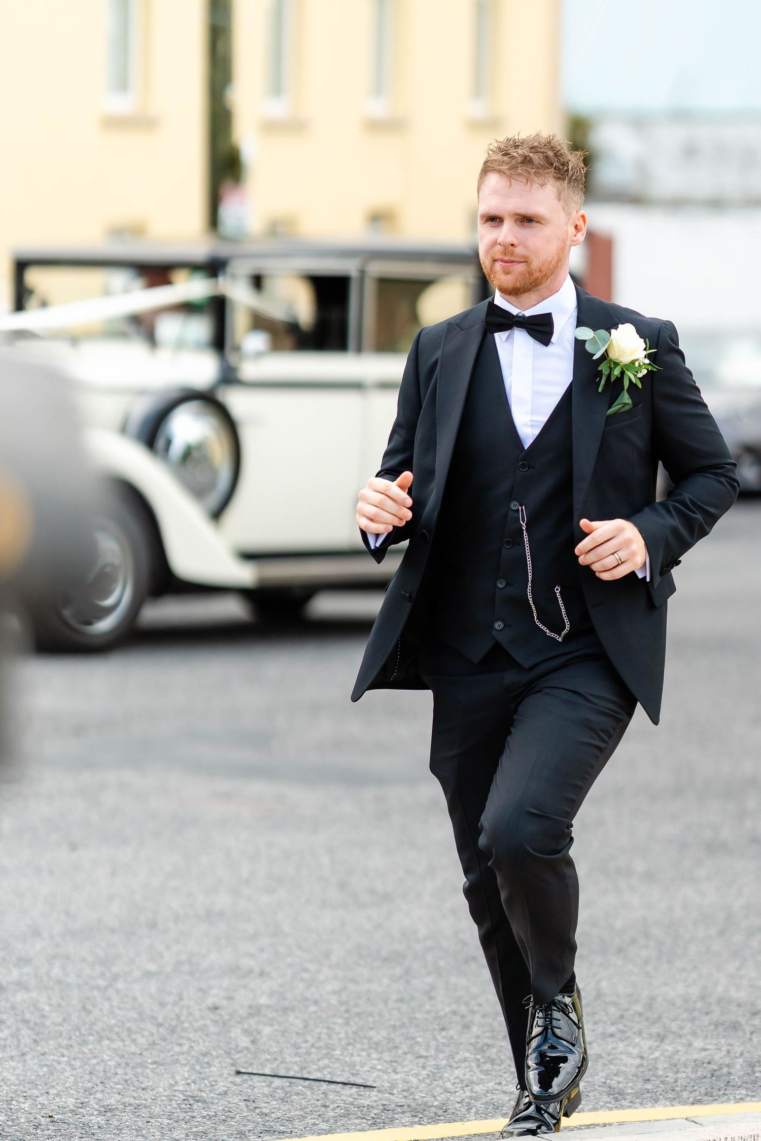 Groom in formal black tuxedo and bowtie walking outdoors near vintage cars.