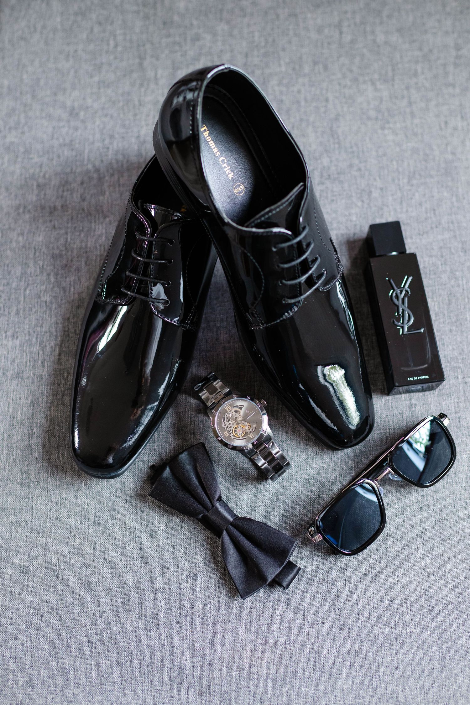Formal black patent leather dress shoes with accessories on a gray textile background.