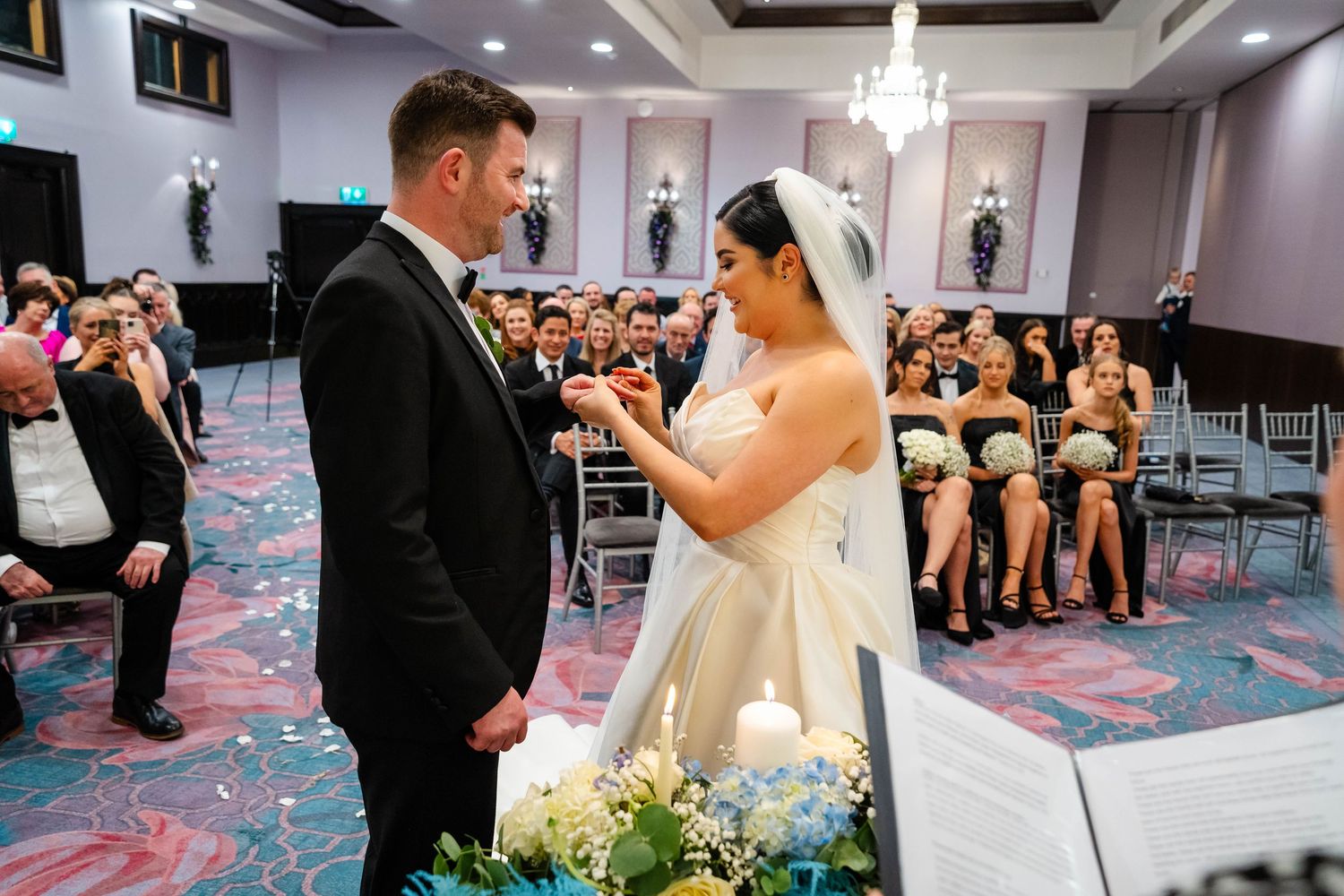 Wedding ceremony in progress with guests seated in a modern ballroom with teal carpet.