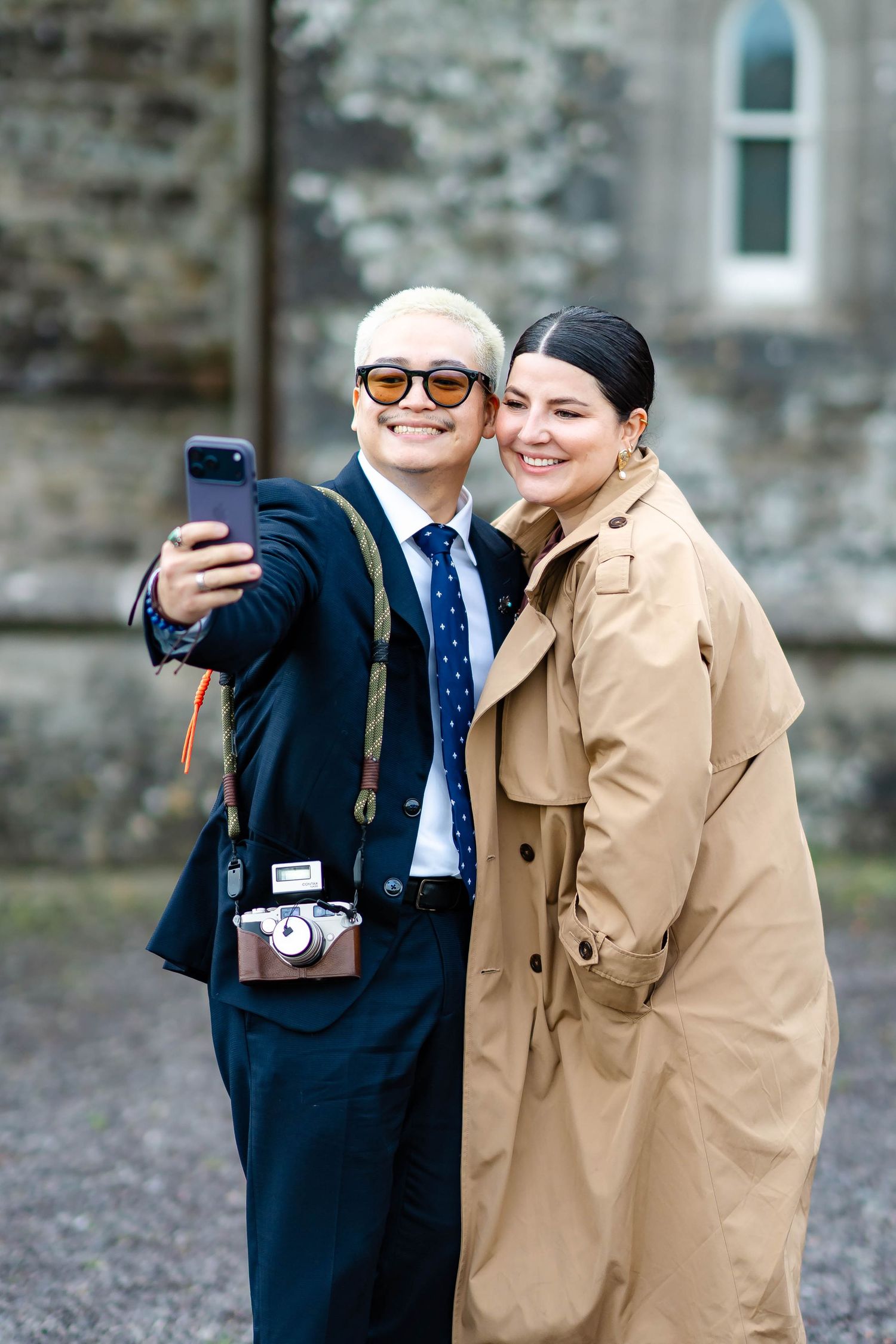 Two people take selfies together outdoors, one wearing a navy suit and one in a beige coat.