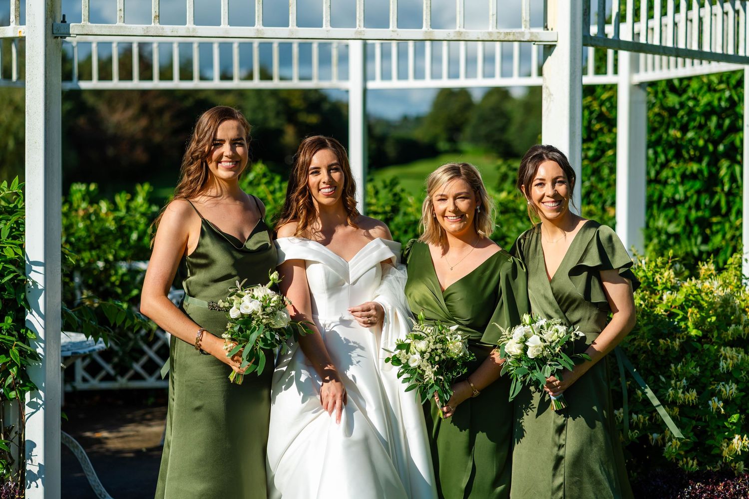 Wedding party in green dresses poses under white pergola with floral decorations.