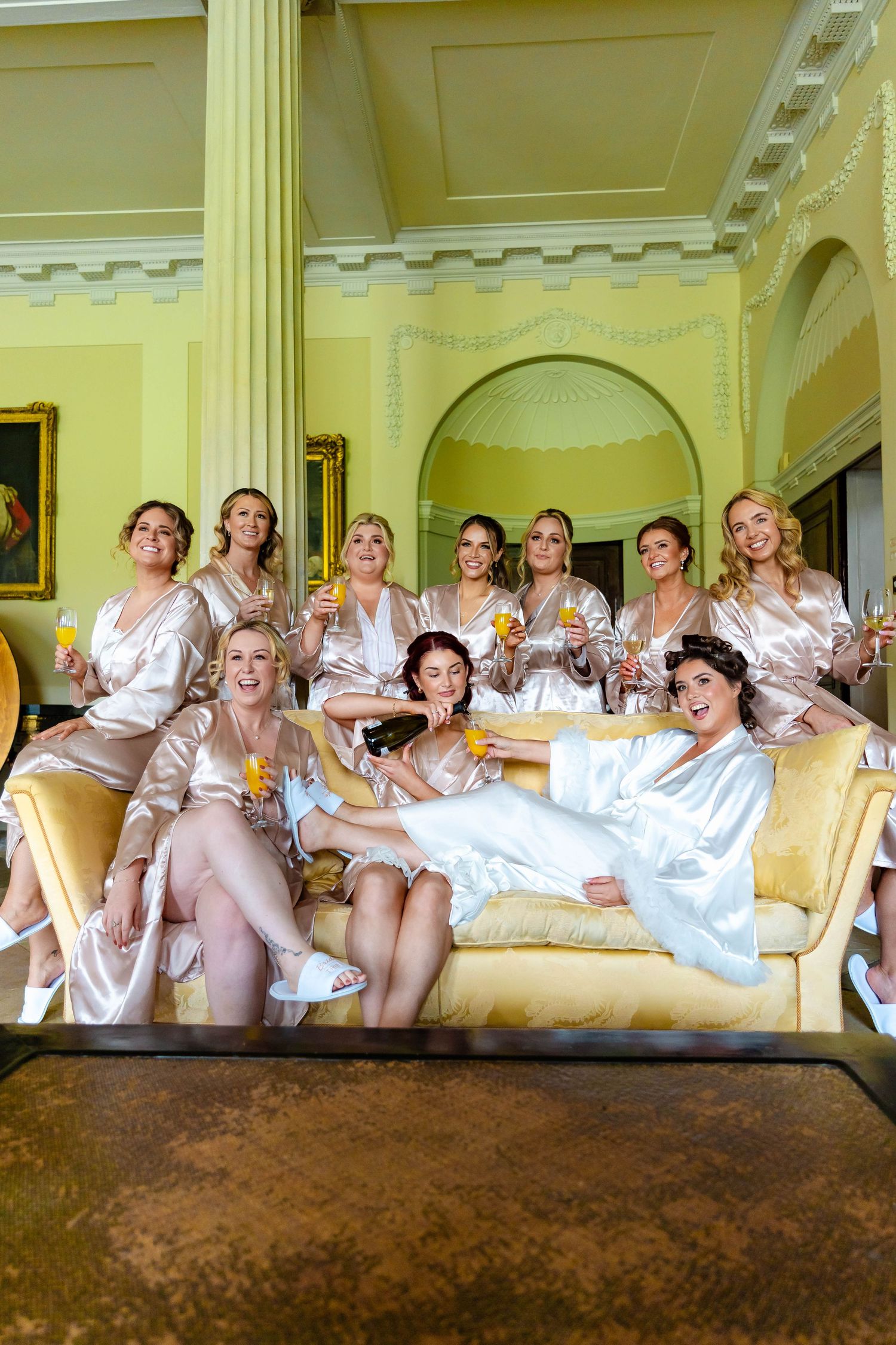 A group of bridesmaids in white robes pose together on an elegant couch in a luxurious room with yellow walls.