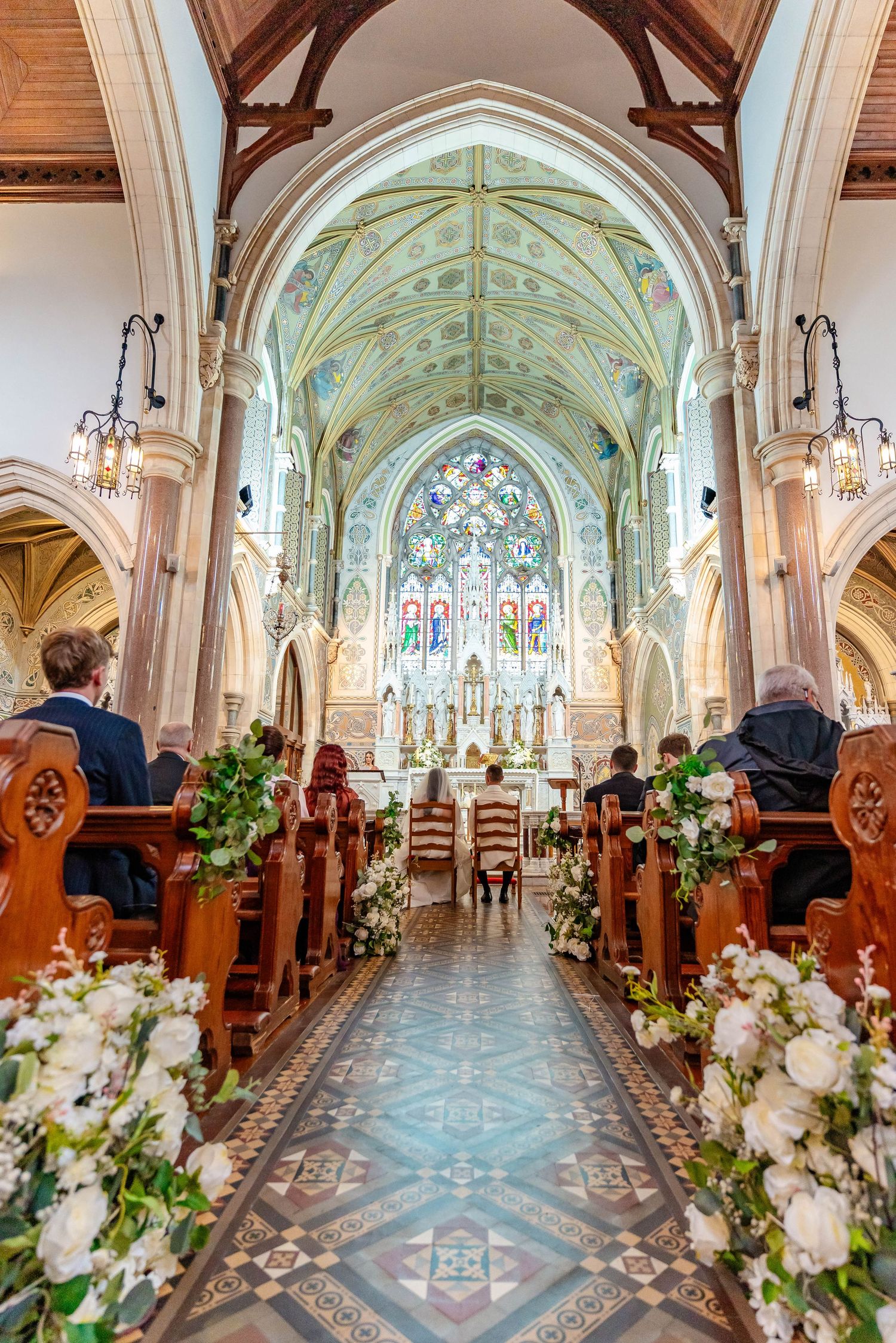 Interior view of a grand cathedral with ornate stained glass windows and floral decorations along the aisle for a wedding ceremony.