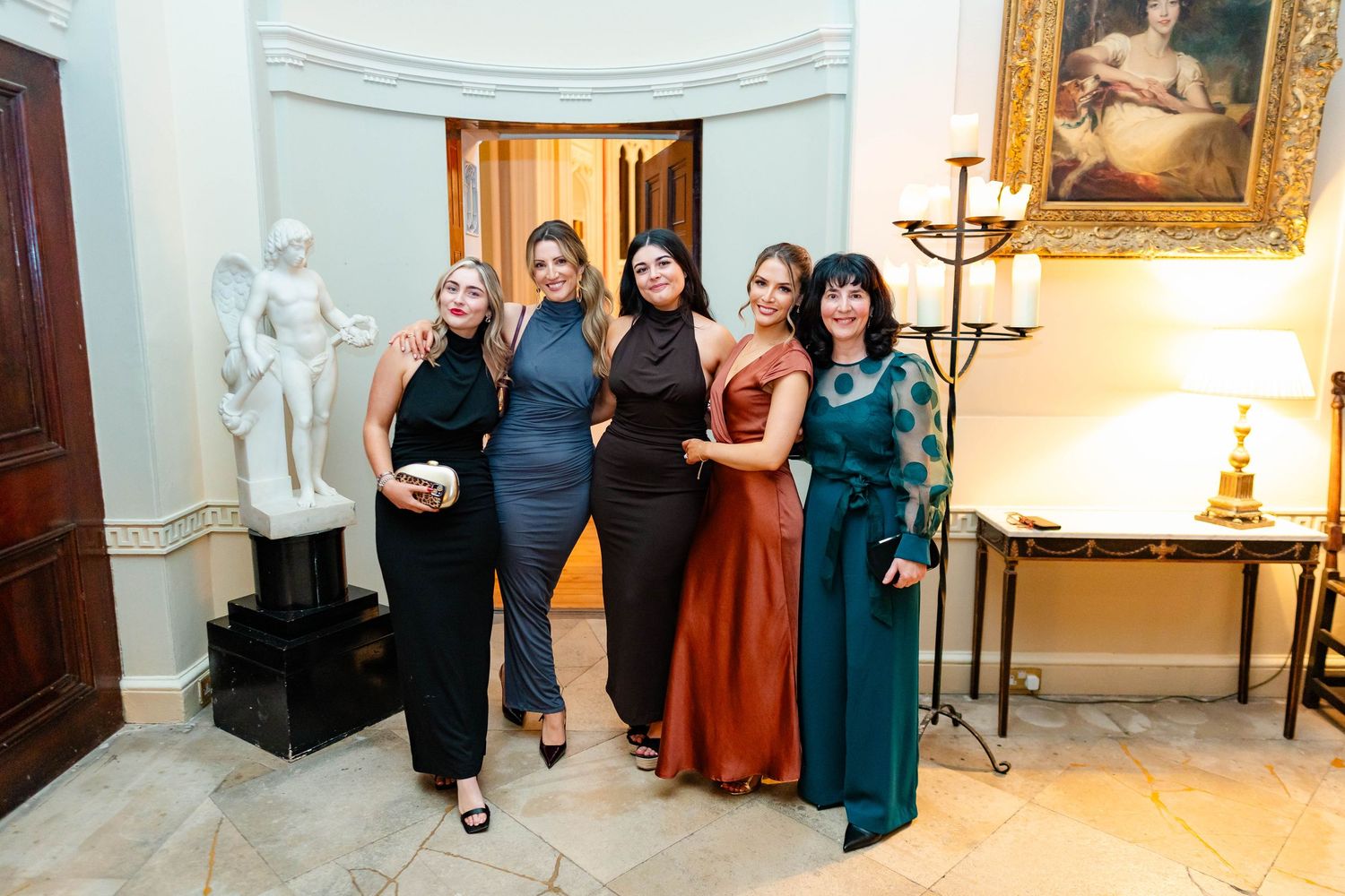 Guests in elegant dresses gather in a grand room with ornate decor and classical paintings on display.