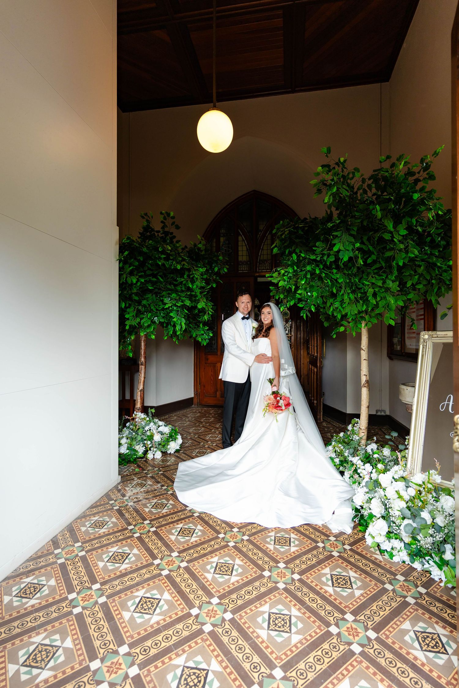 A couple poses on a tiled patio with white columns and green plants creating a romantic wedding scene.