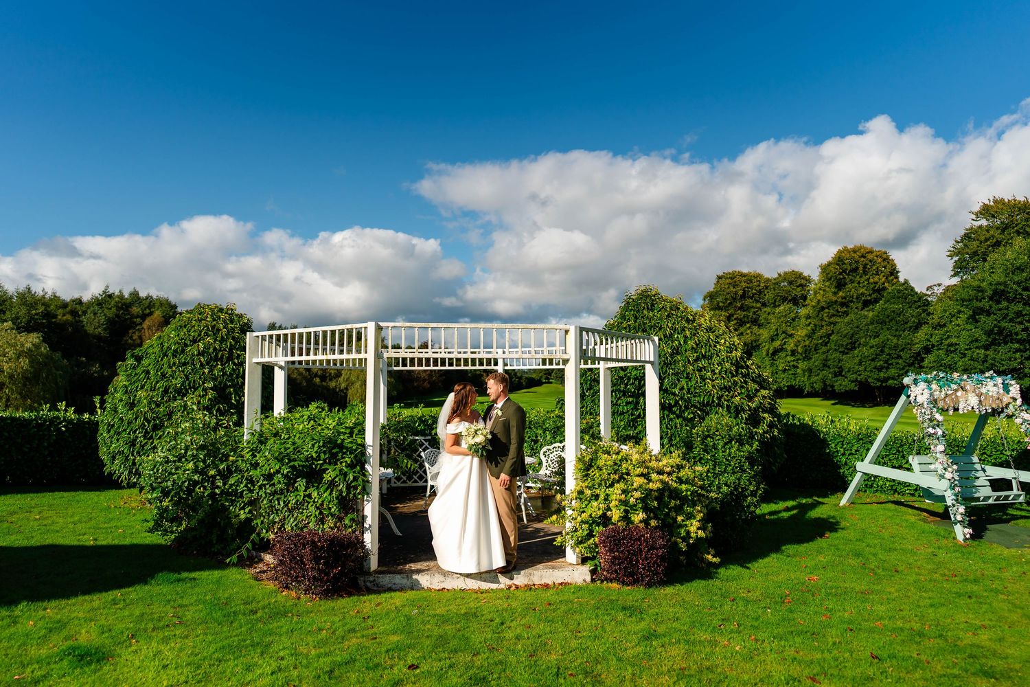 Wedding ceremony setup with white pergola and chairs on a lush green lawn overlooking gardens on a sunny day.
