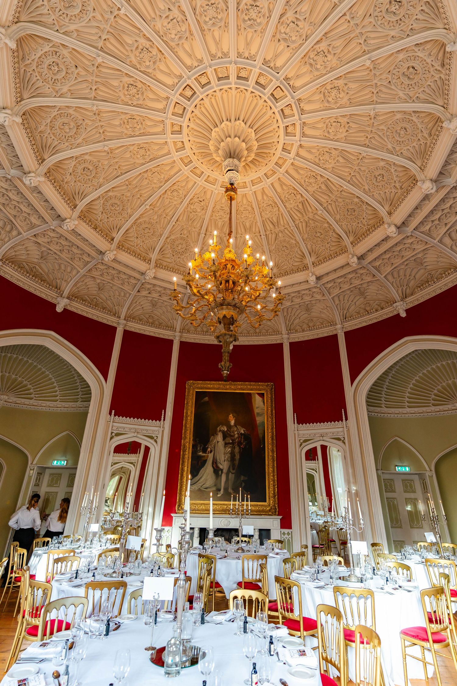 Ornate white and gold ceiling with chandeliers in an elegant dining room featuring red walls and formal table settings.