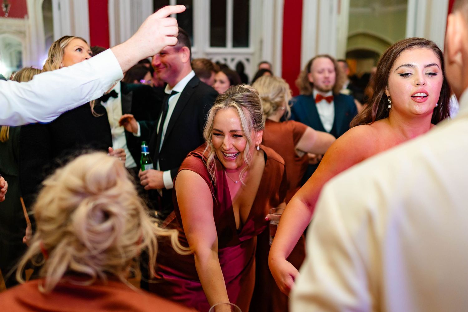 Energetic crowd dancing and celebrating at an indoor wedding reception with decorative lighting.