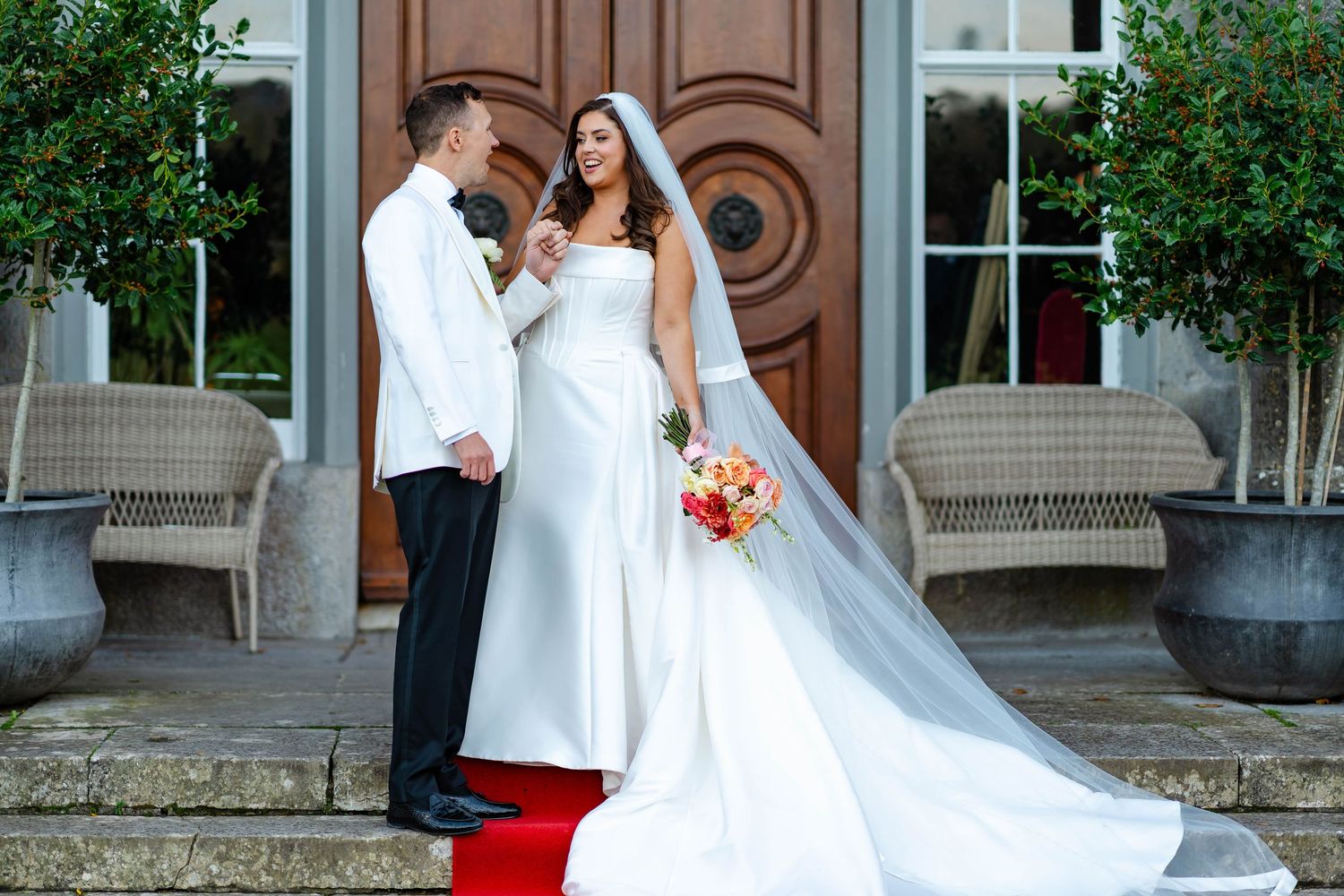 A couple in formal wedding attire poses on stone steps outside an elegant building with ornate wooden doors.