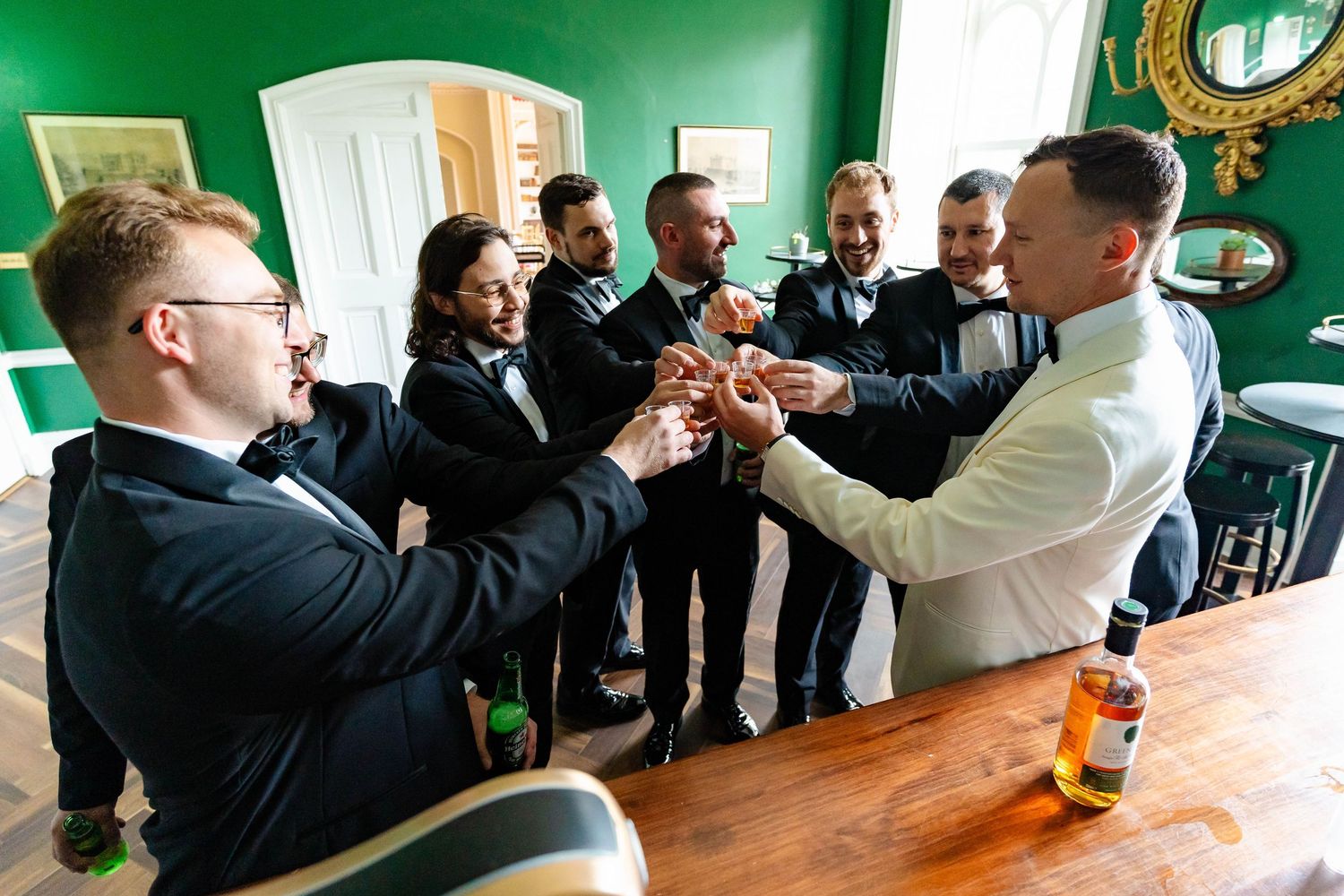 Groomsmen in black suits gather around a bar counter for drinks and conversation at a wedding reception.