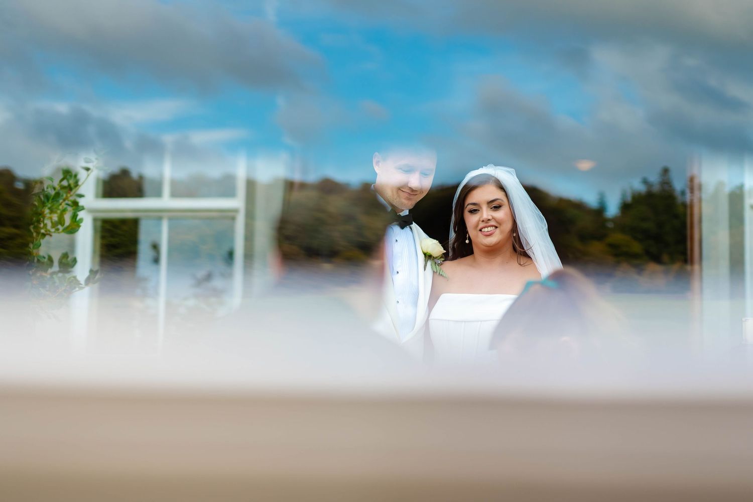 A wedding photo captured through a creative blurred foreground framing with dreamy clouds in the background.