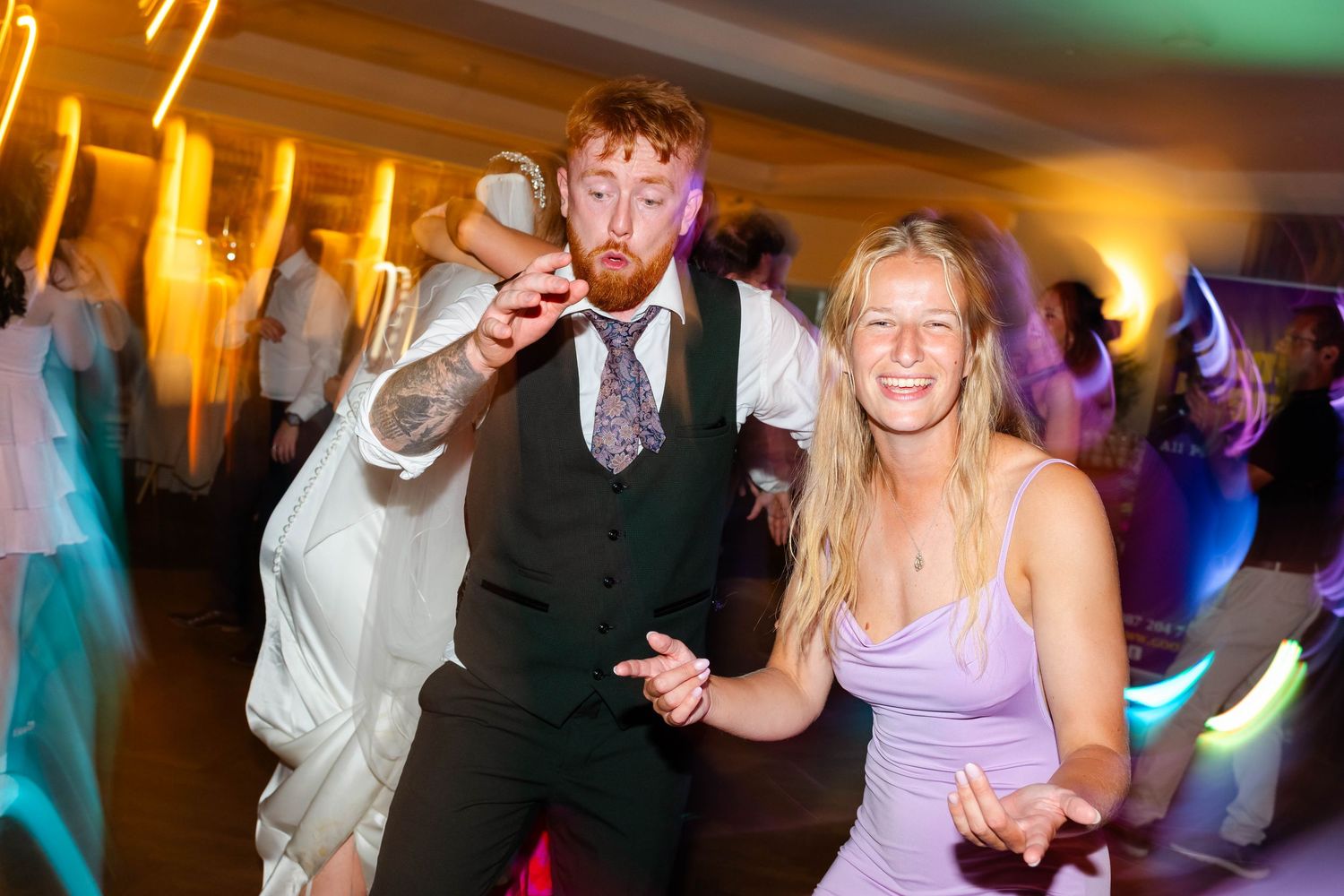Wedding guests dancing energetically on a colorful dance floor at an indoor reception venue.