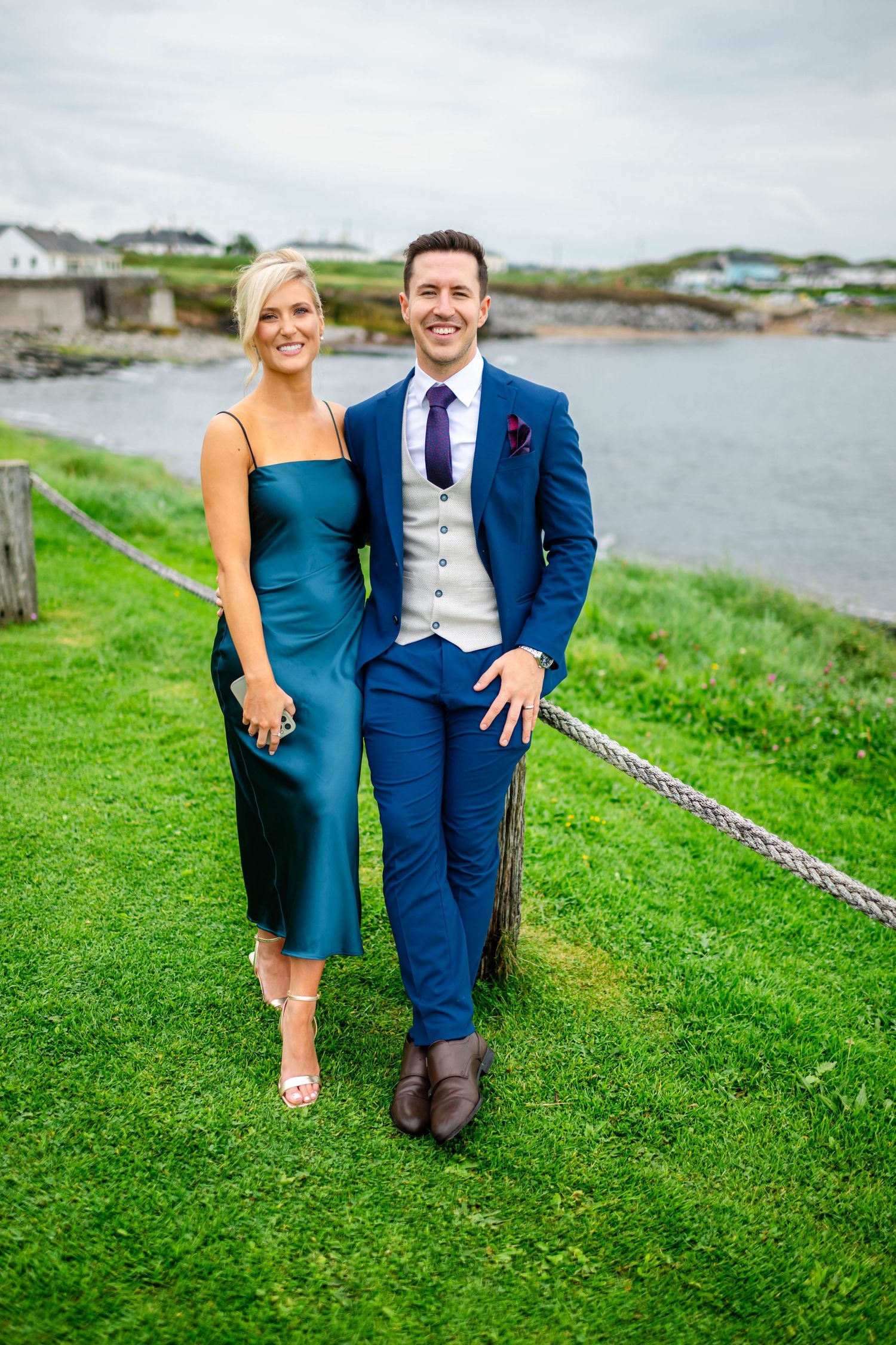 Couples in formal attire pose on grassy hillside overlooking coastal waters.