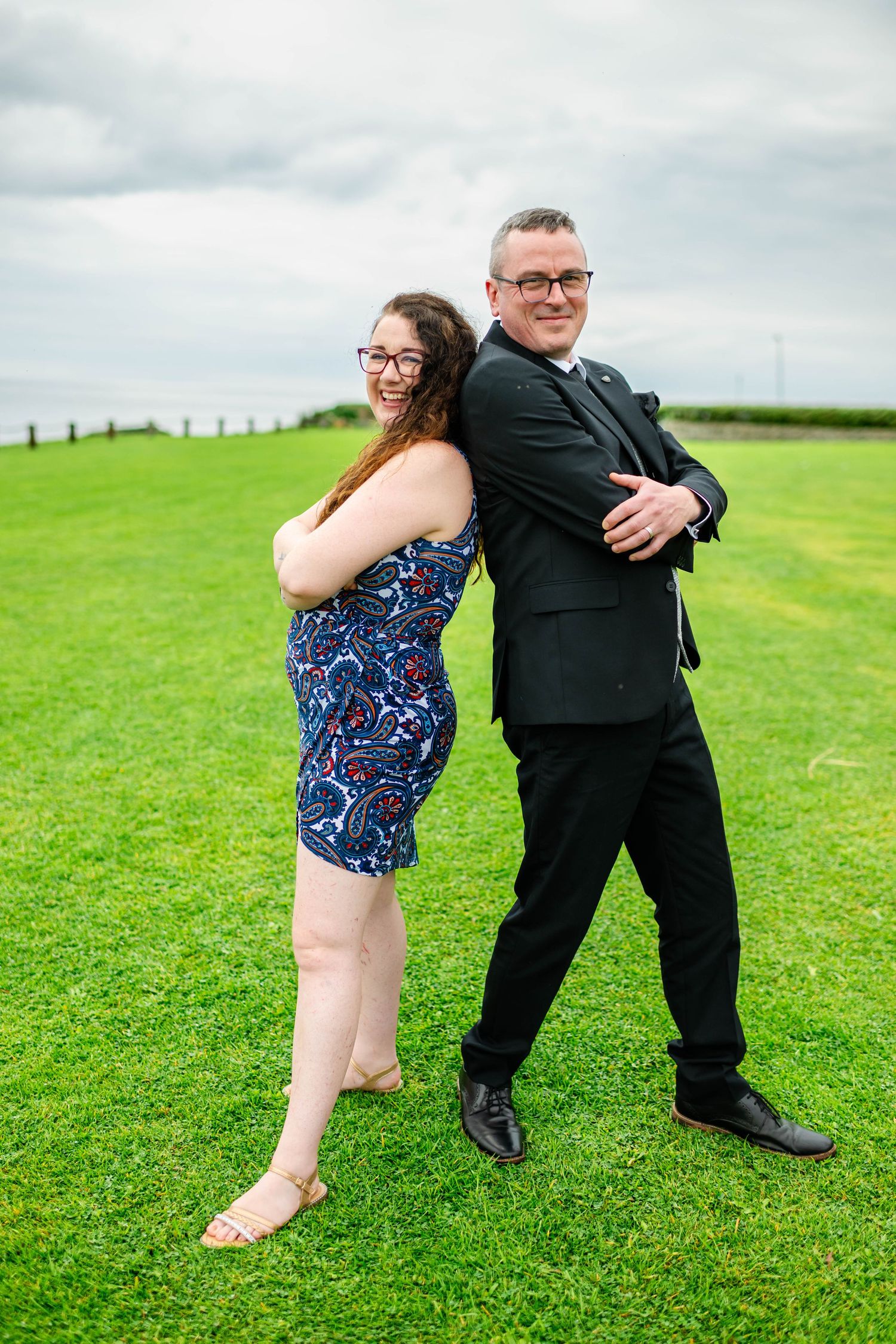 Playful couple in formal wear dance on green grass during outdoor celebration.