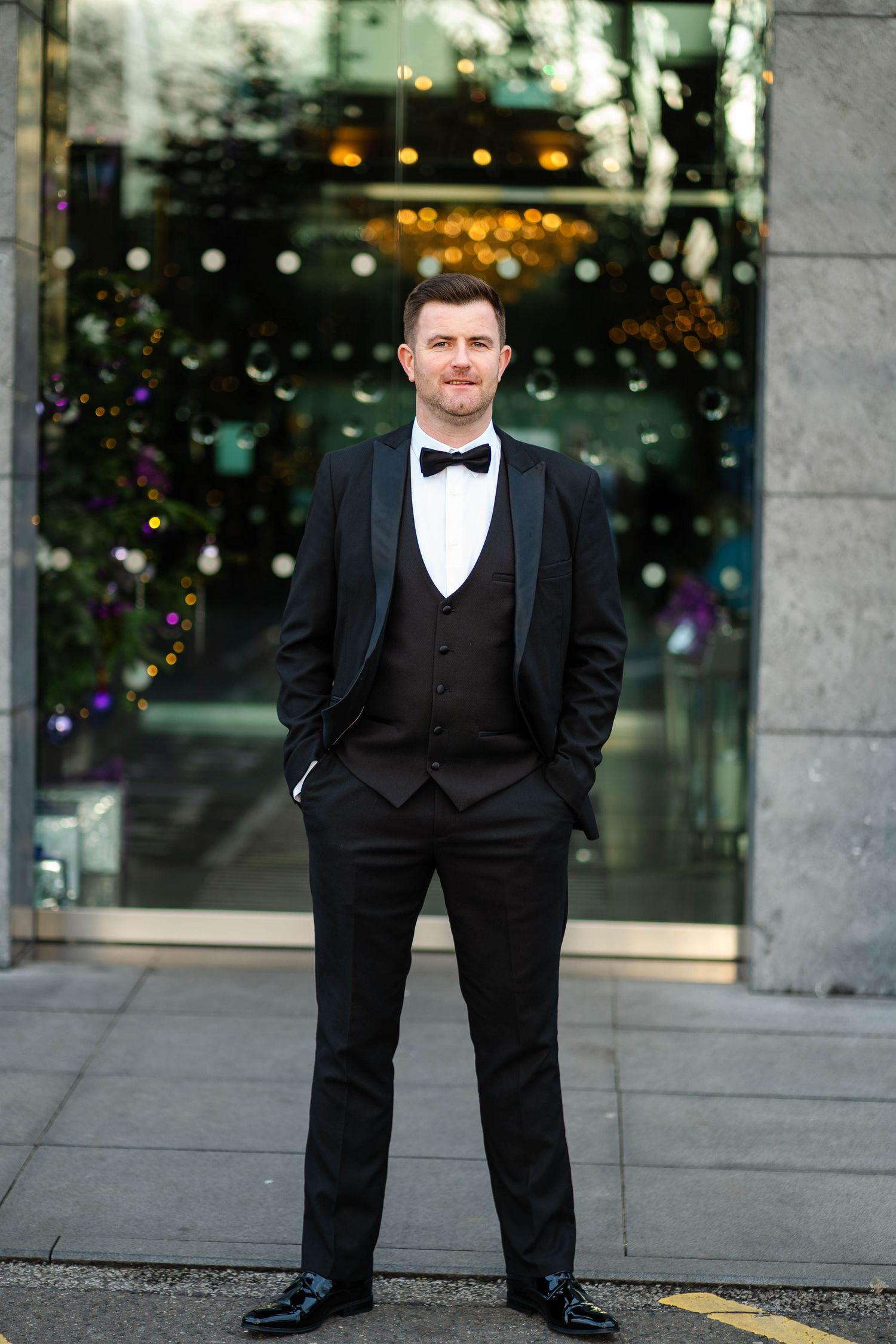 Person in formal black tuxedo standing outside a building with decorative lights.