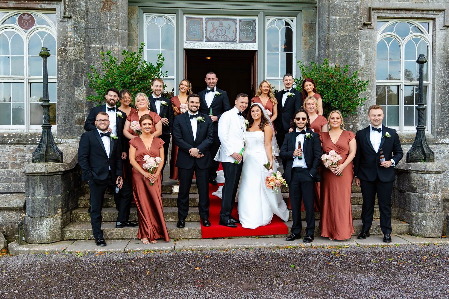 Wedding party poses on stone steps of elegant mansion with red carpet and gothic windows in background.