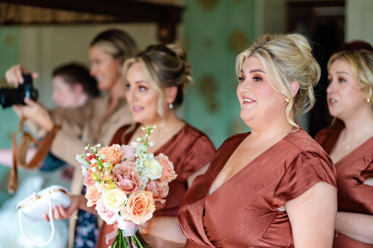 Bridesmaids in burgundy dresses stand in a row holding peach and pink floral bouquets during a wedding ceremony.
