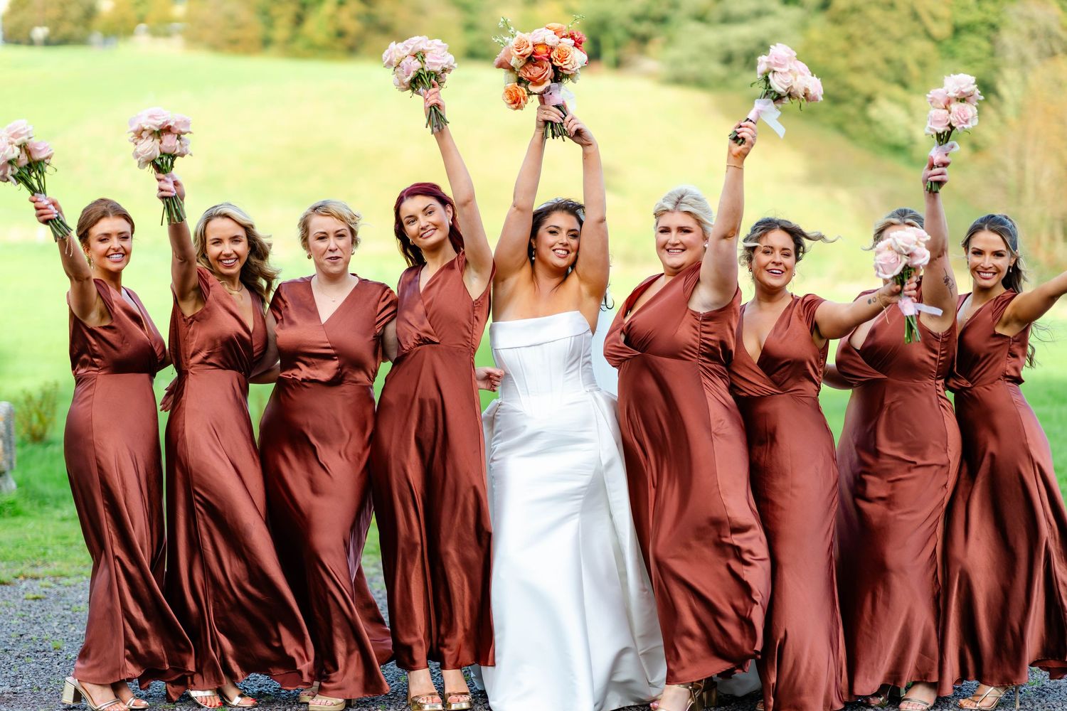 A bridal party in flowing rust-colored dresses and one white wedding gown pose joyfully with bouquets on a grassy lawn.