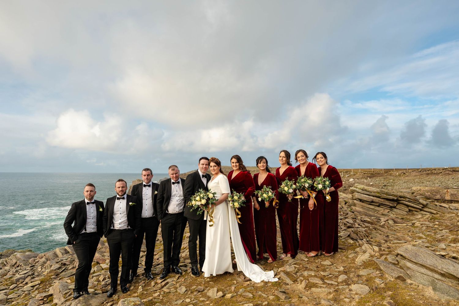 Wedding party in burgundy and black attire poses on rocky coastal cliff with ocean backdrop.