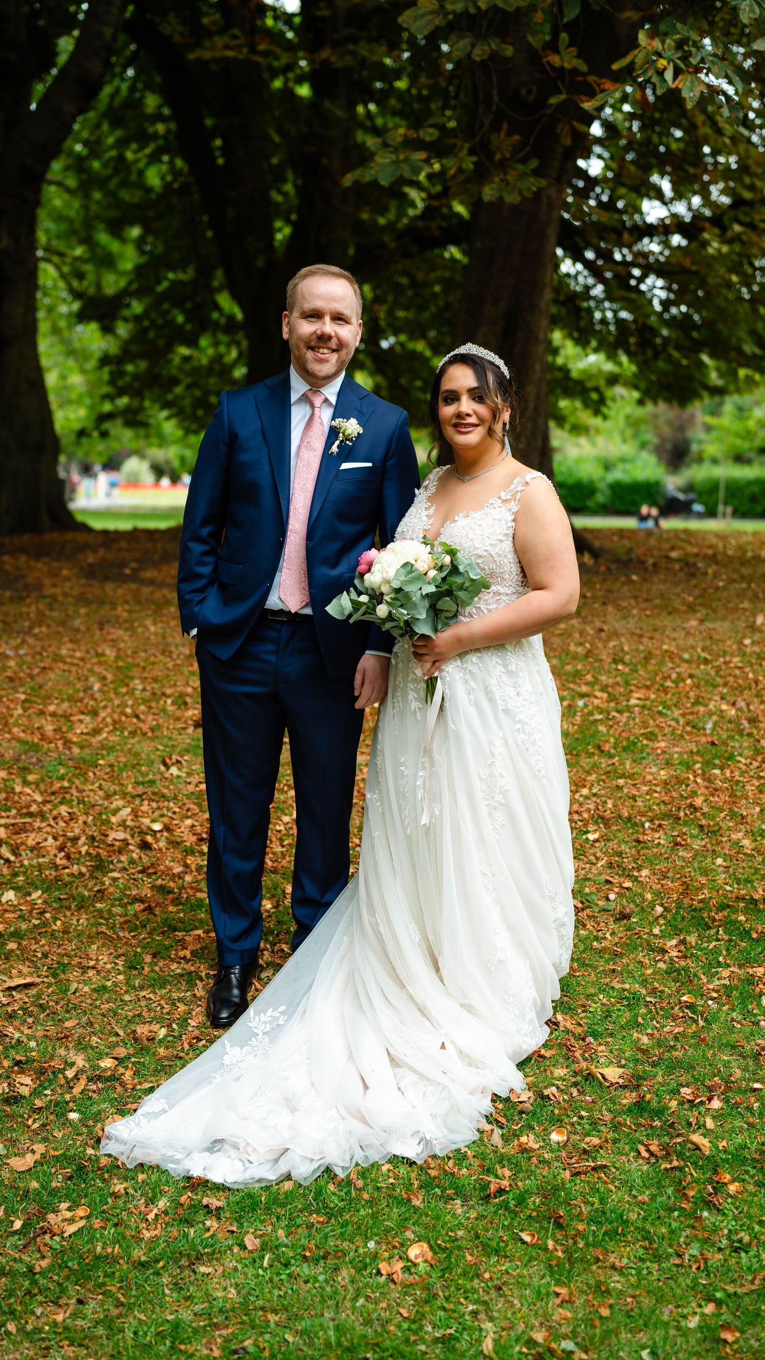 A wedding couple poses together on grass covered with fallen autumn leaves.