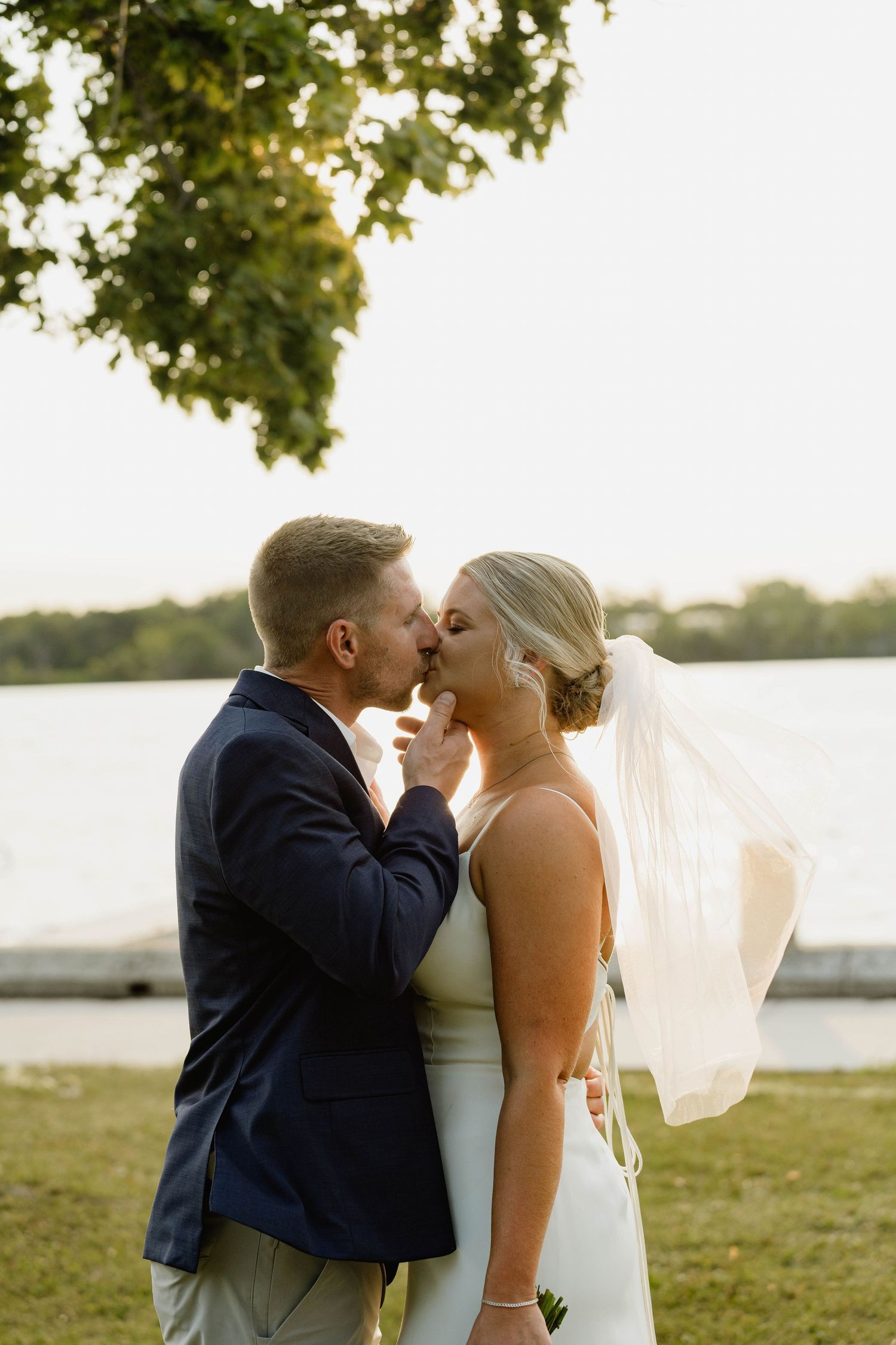 Bride and groom share a romantic kiss outdoors near a lake at sunset, with the bride wearing a white gown and veil.
