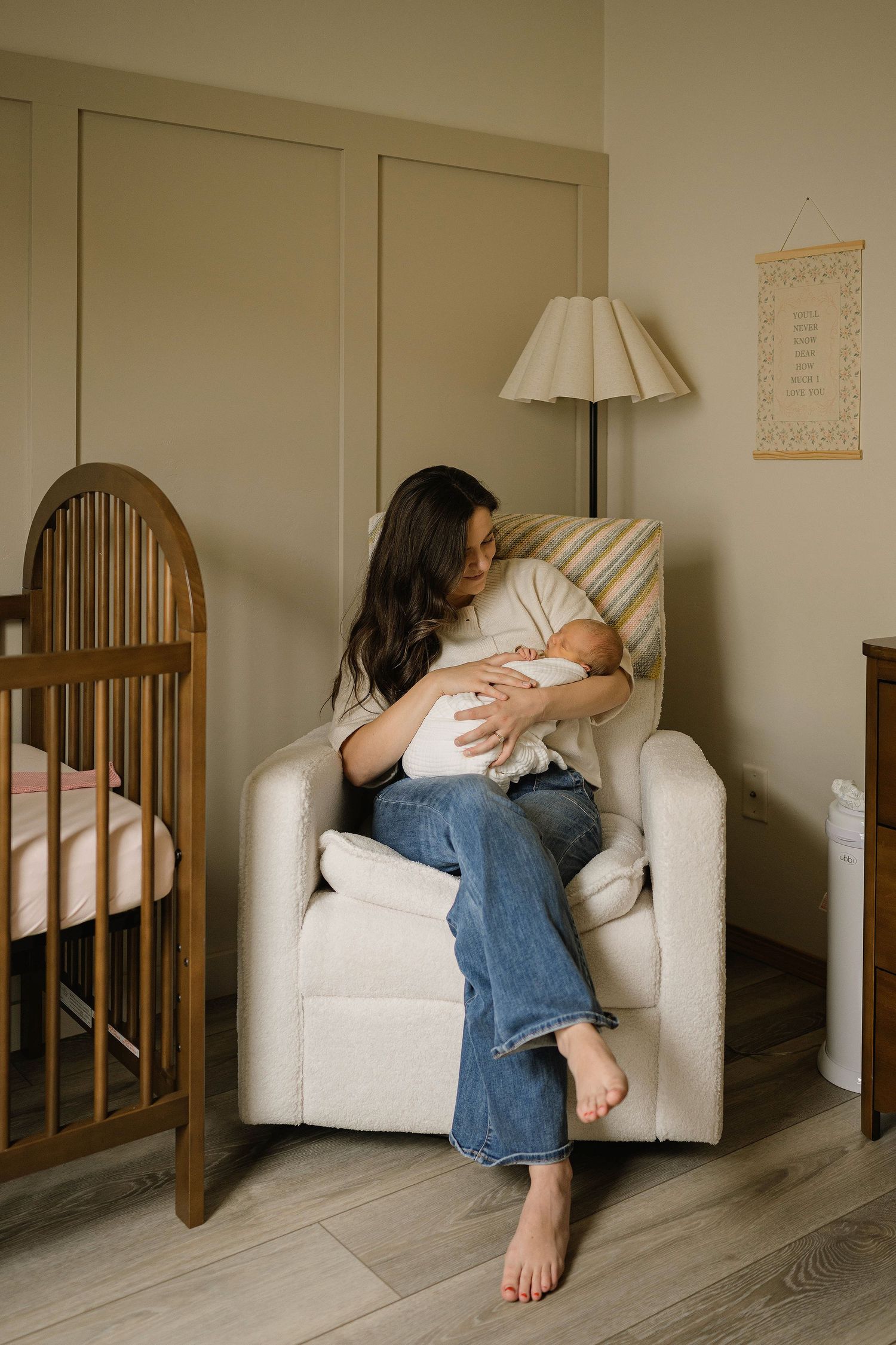 Mother sitting in nursery glider chair holding and bonding with newborn baby near wooden crib.